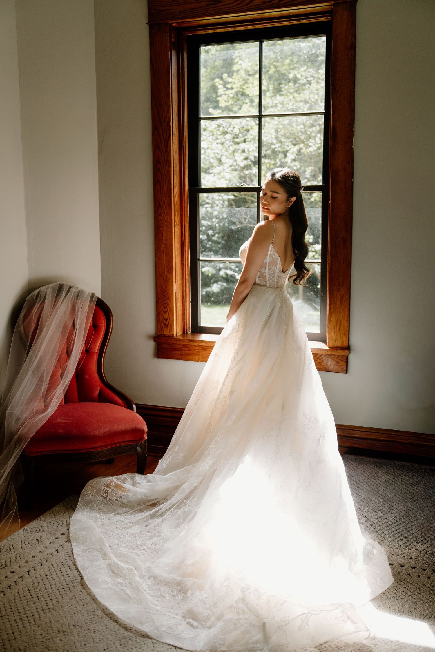 A bride in a flowing white wedding gown stands by a window with natural light streaming in, next to a red velvet chair.