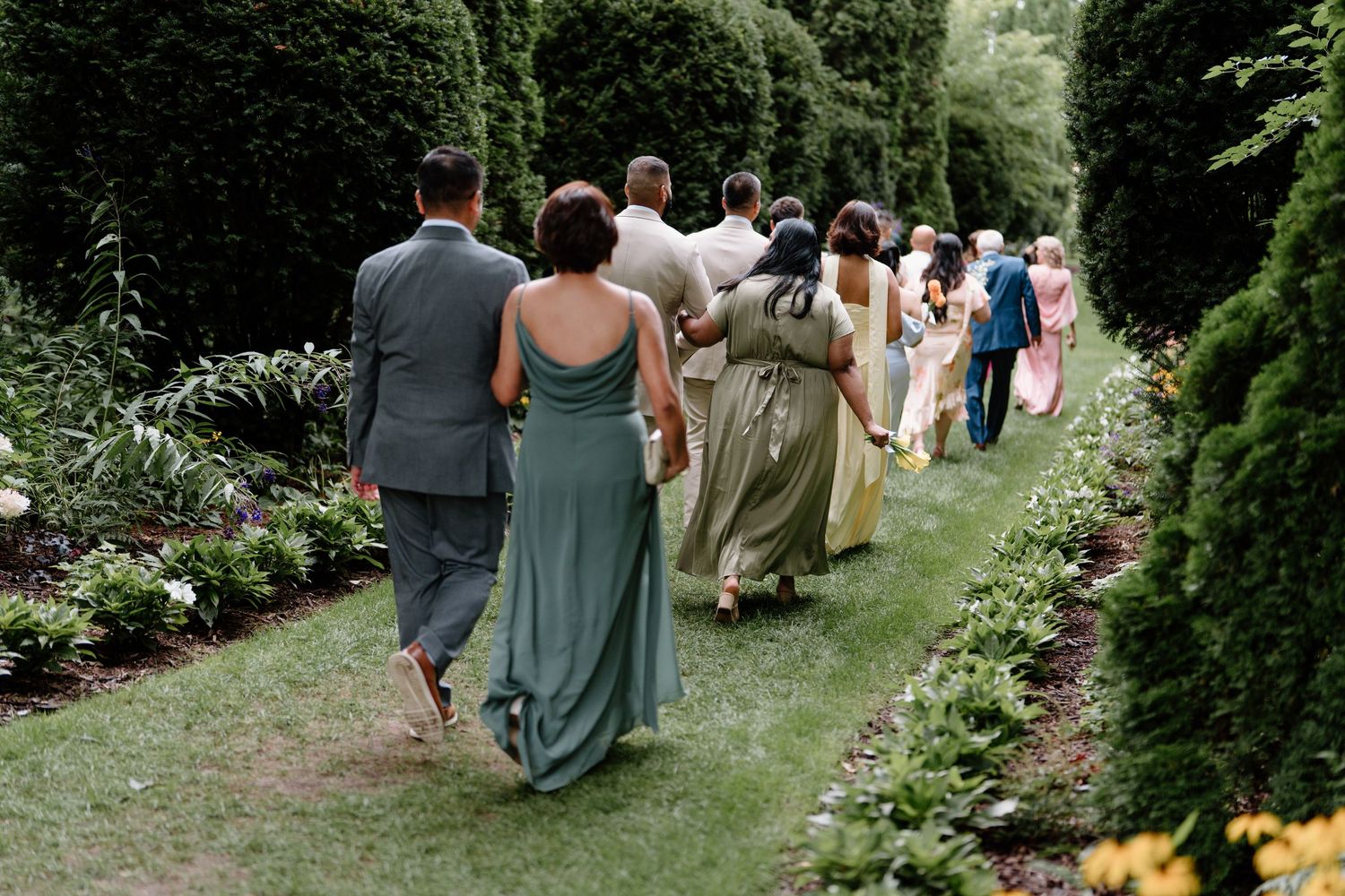 Wedding party walks in a line along a garden path surrounded by lush greenery and flowers.