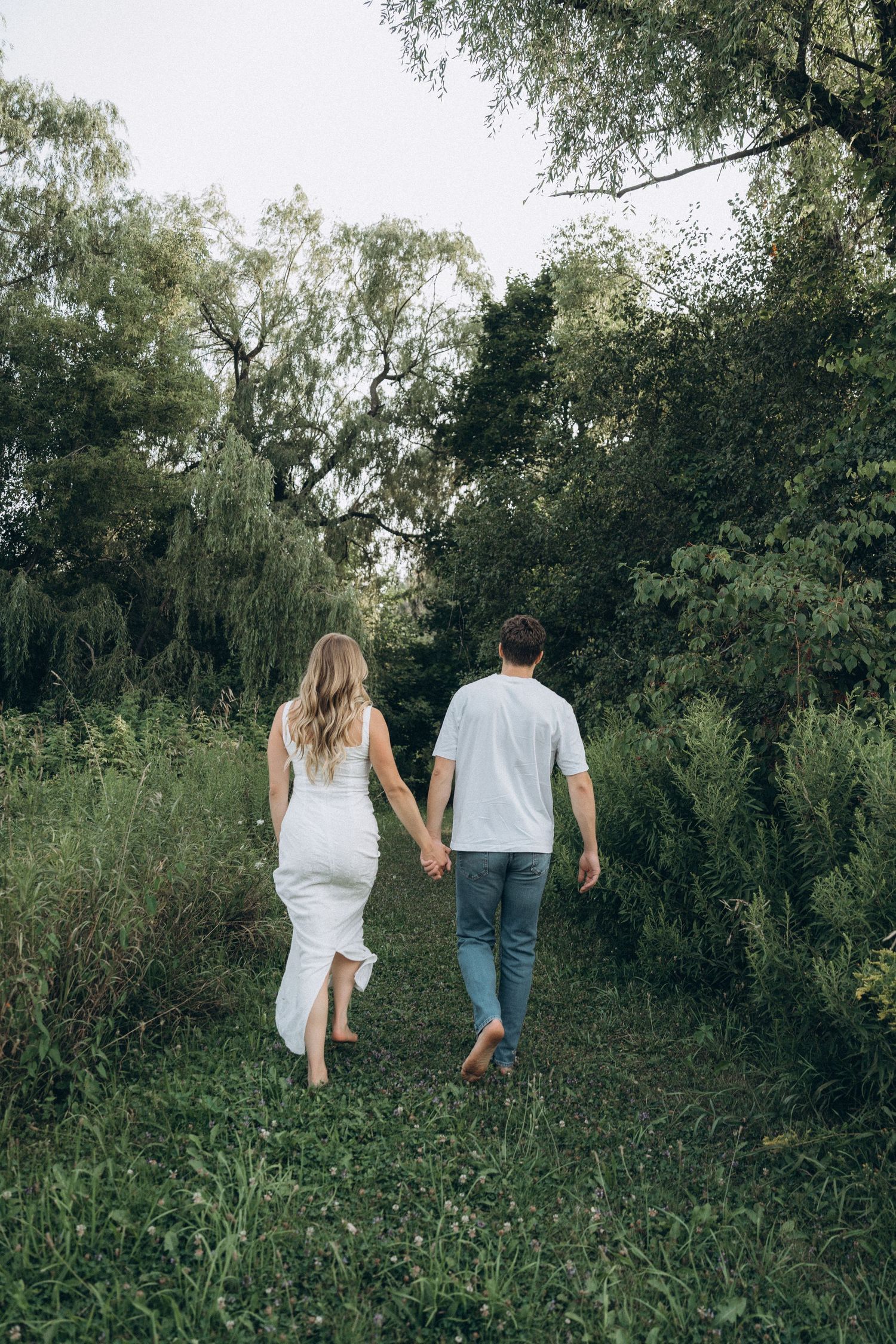A couple walks hand in hand through a lush green path surrounded by trees and tall grass on a summer day.