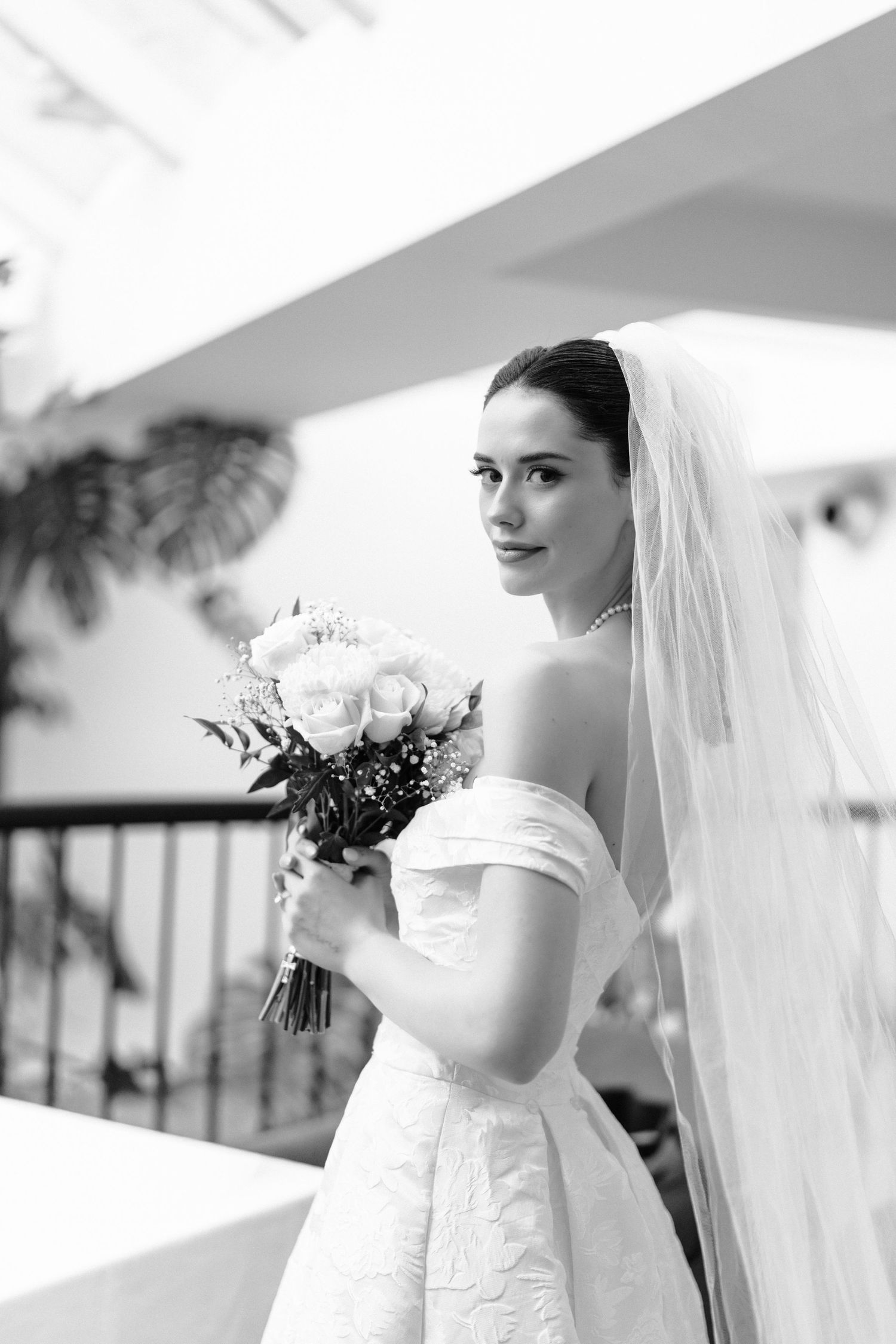 A bride in an off-shoulder wedding gown holds a white rose bouquet while looking over her shoulder in black and white.