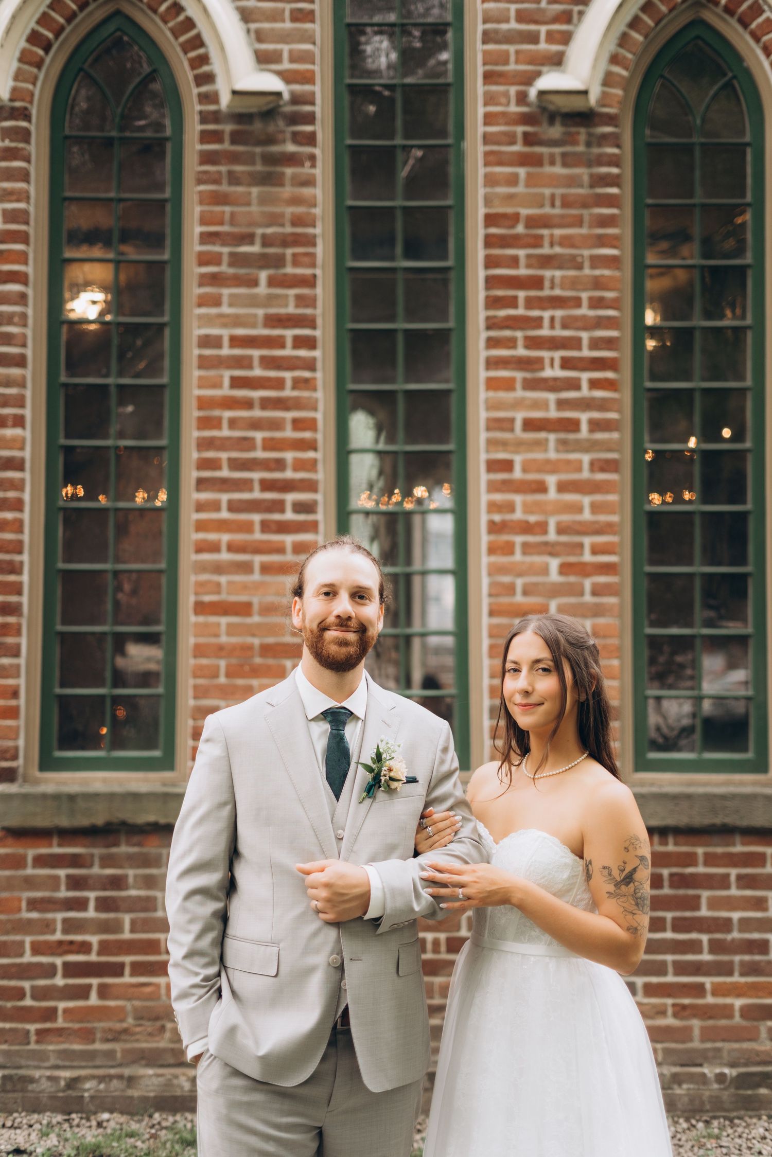 Wedding party in formal attire poses outside historic brick church with arched windows.