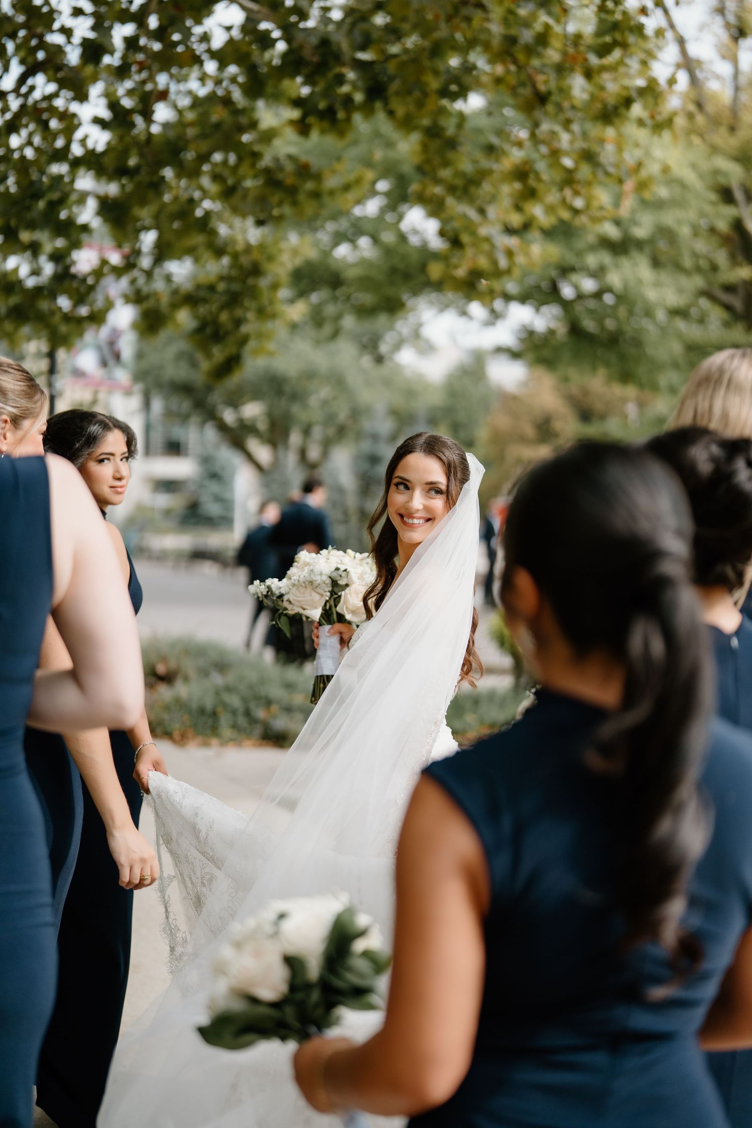 Wedding party in navy and white attire gather outdoors under trees for formal photos on a sunny day.
