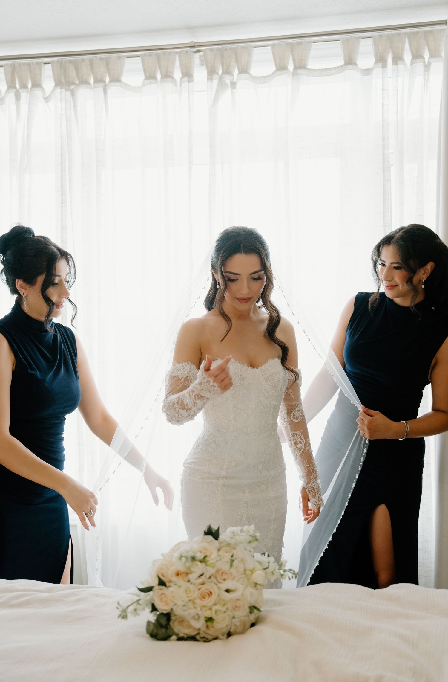 Wedding party members in navy blue dresses help adjust a bride's white gown in a bright sunlit room.