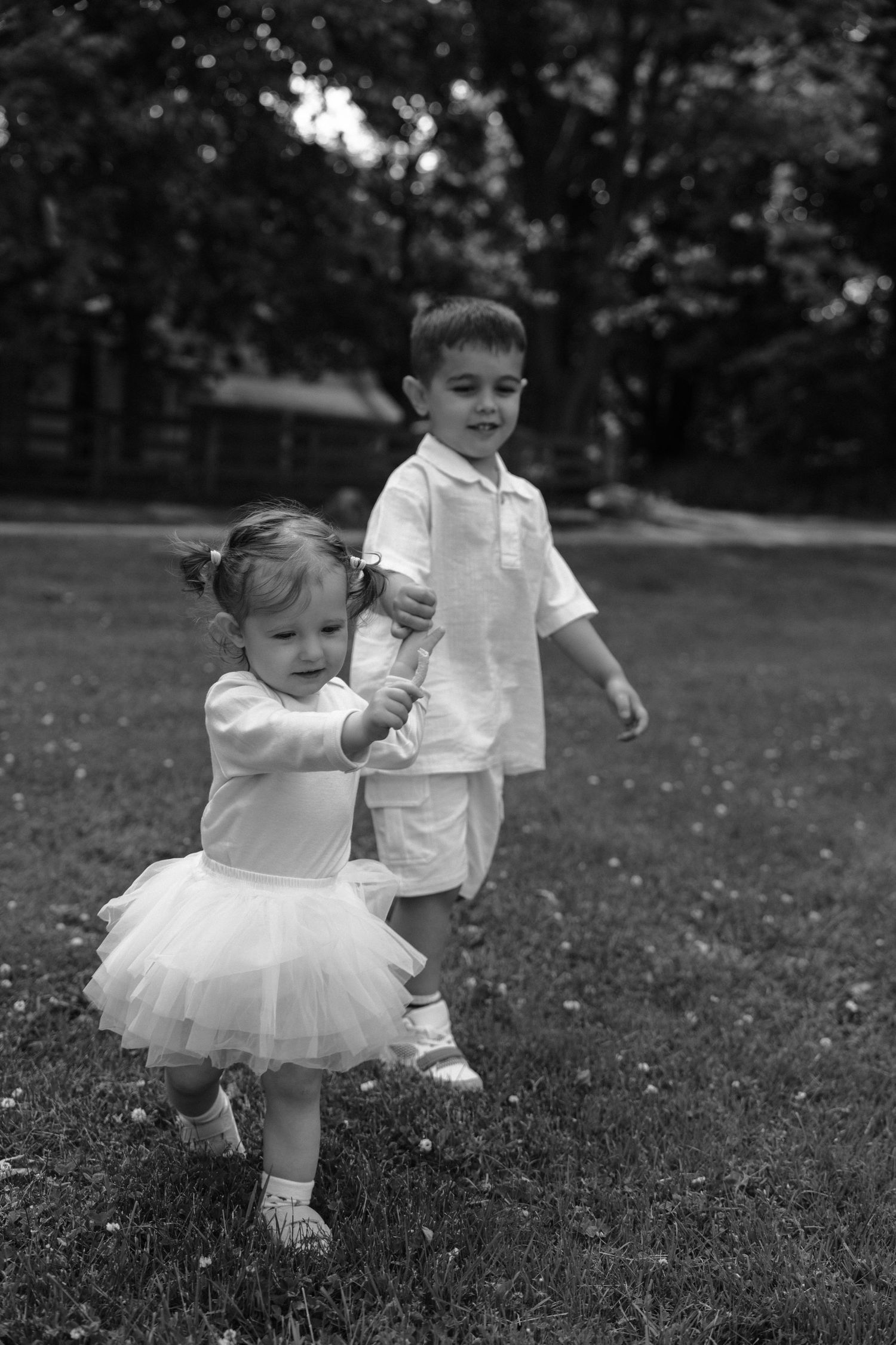 Two children playing in a garden, with a toddler wearing a white tutu and holding hands with sibling in white shirt.