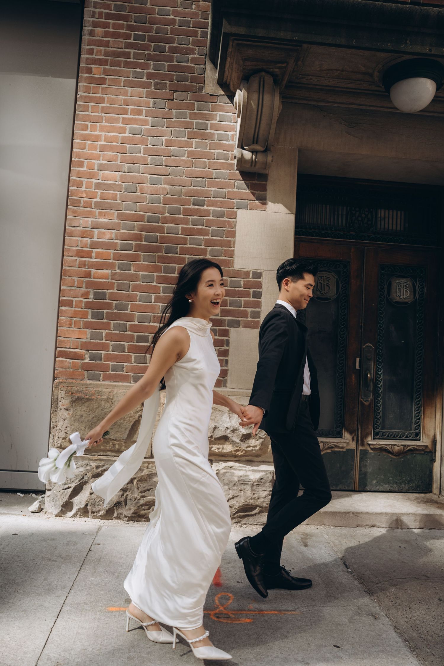 A couple in wedding attire holds hands while walking along a sidewalk against a brick building facade.