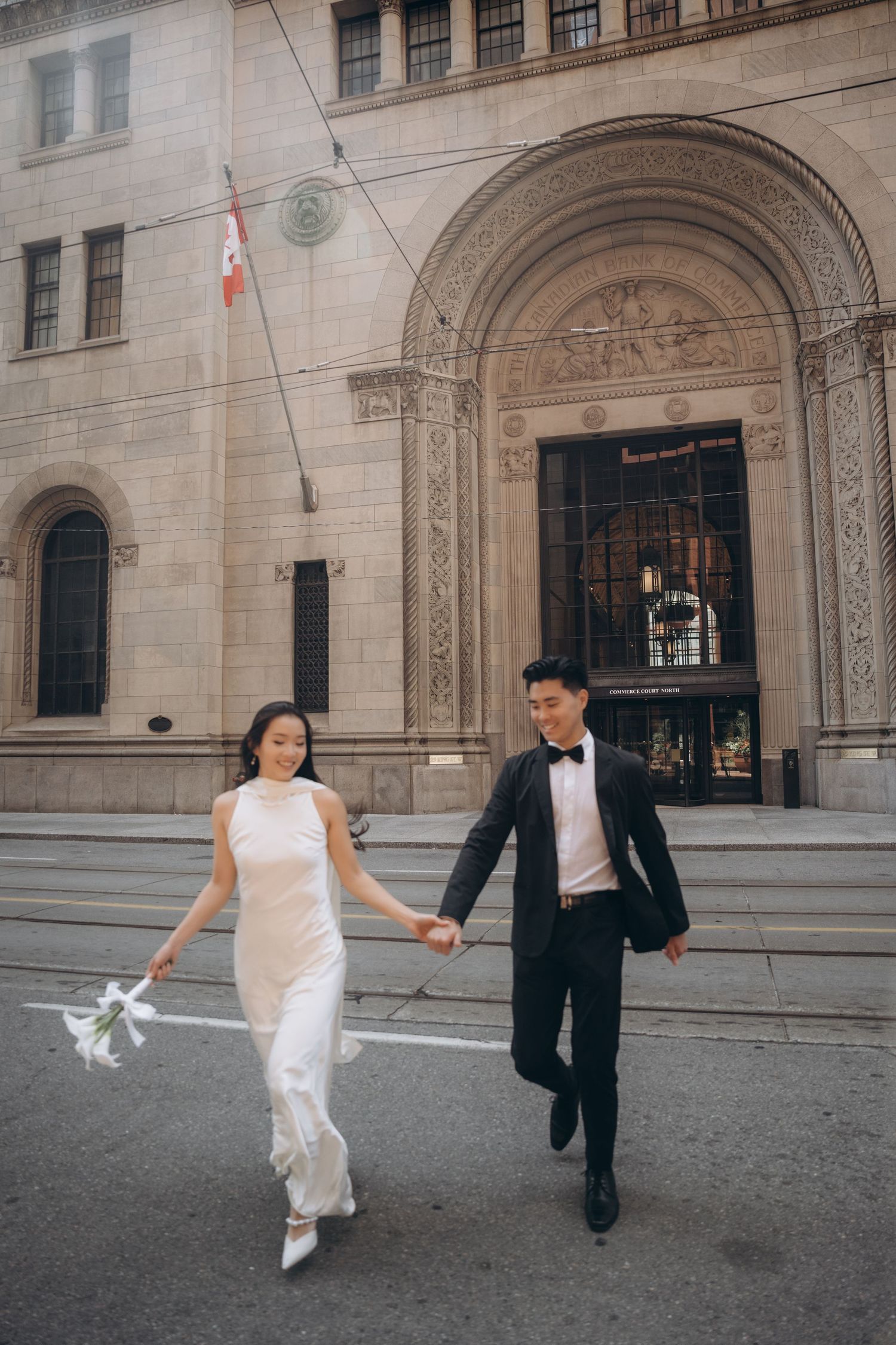 A couple in formal attire runs joyfully together in front of a grand historic building entrance.