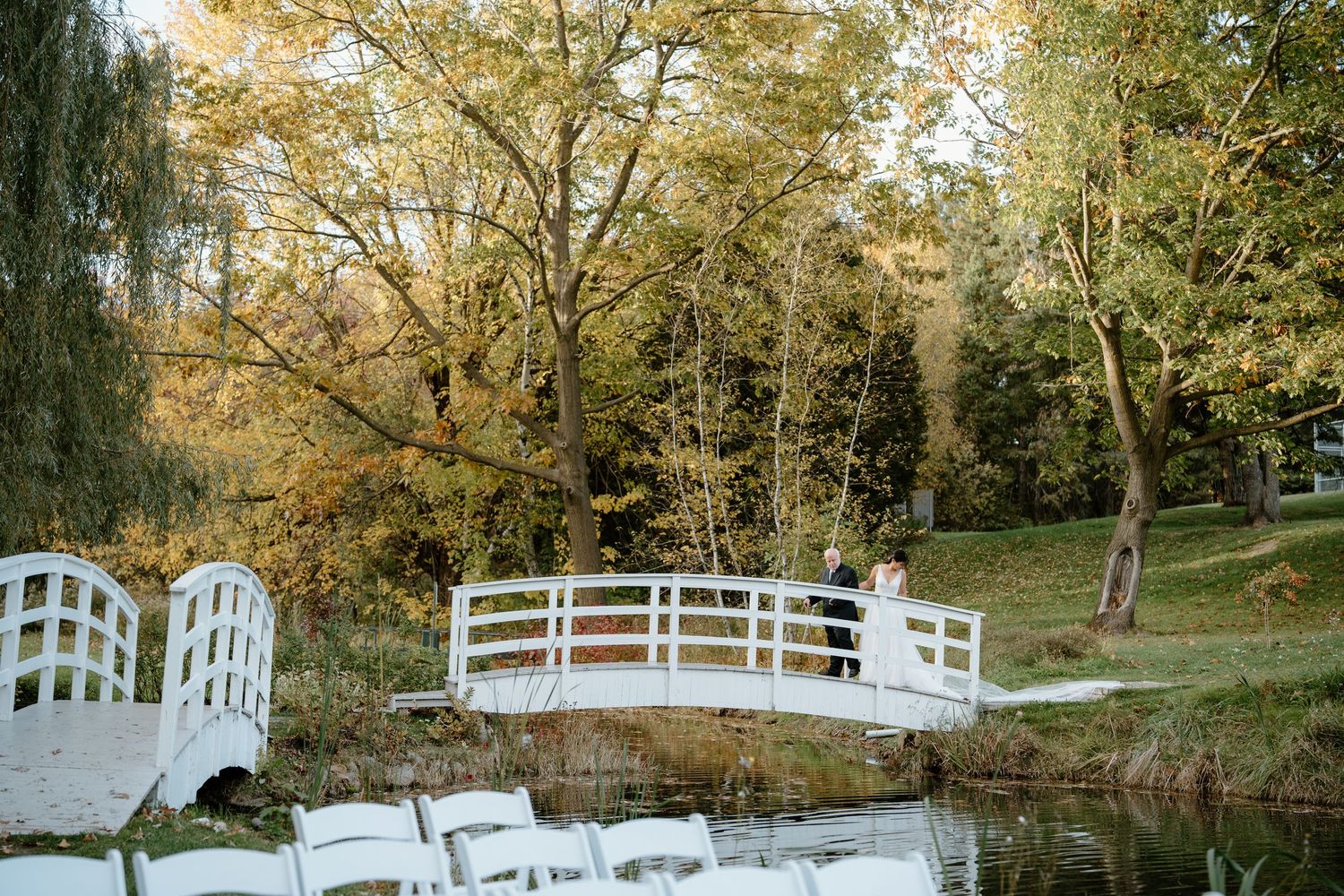 White decorative bridge over water with wedding chairs setup nearby during fall season.