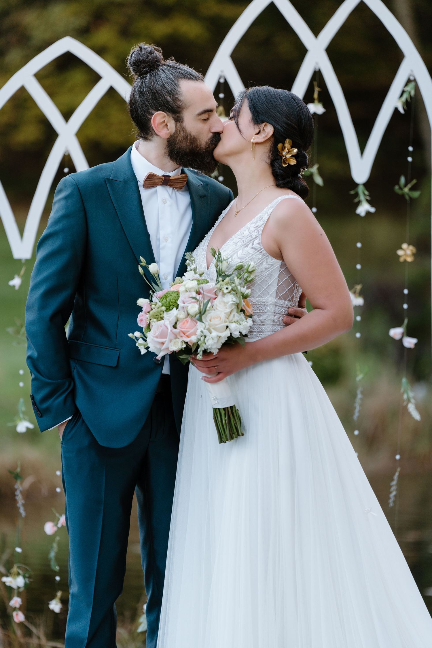Newlyweds share a kiss under a white arch, with the bride holding a delicate pastel floral bouquet.