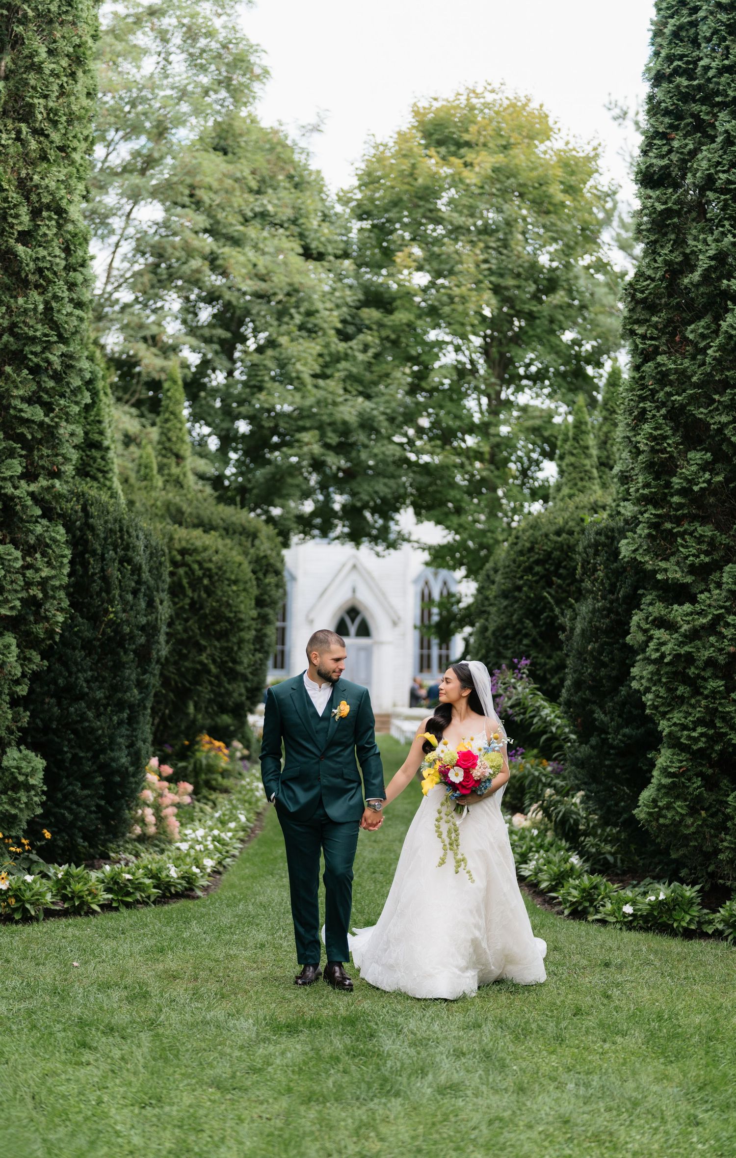 Wedding couple walks together through a lush garden setting with green trees and white chapel in background.