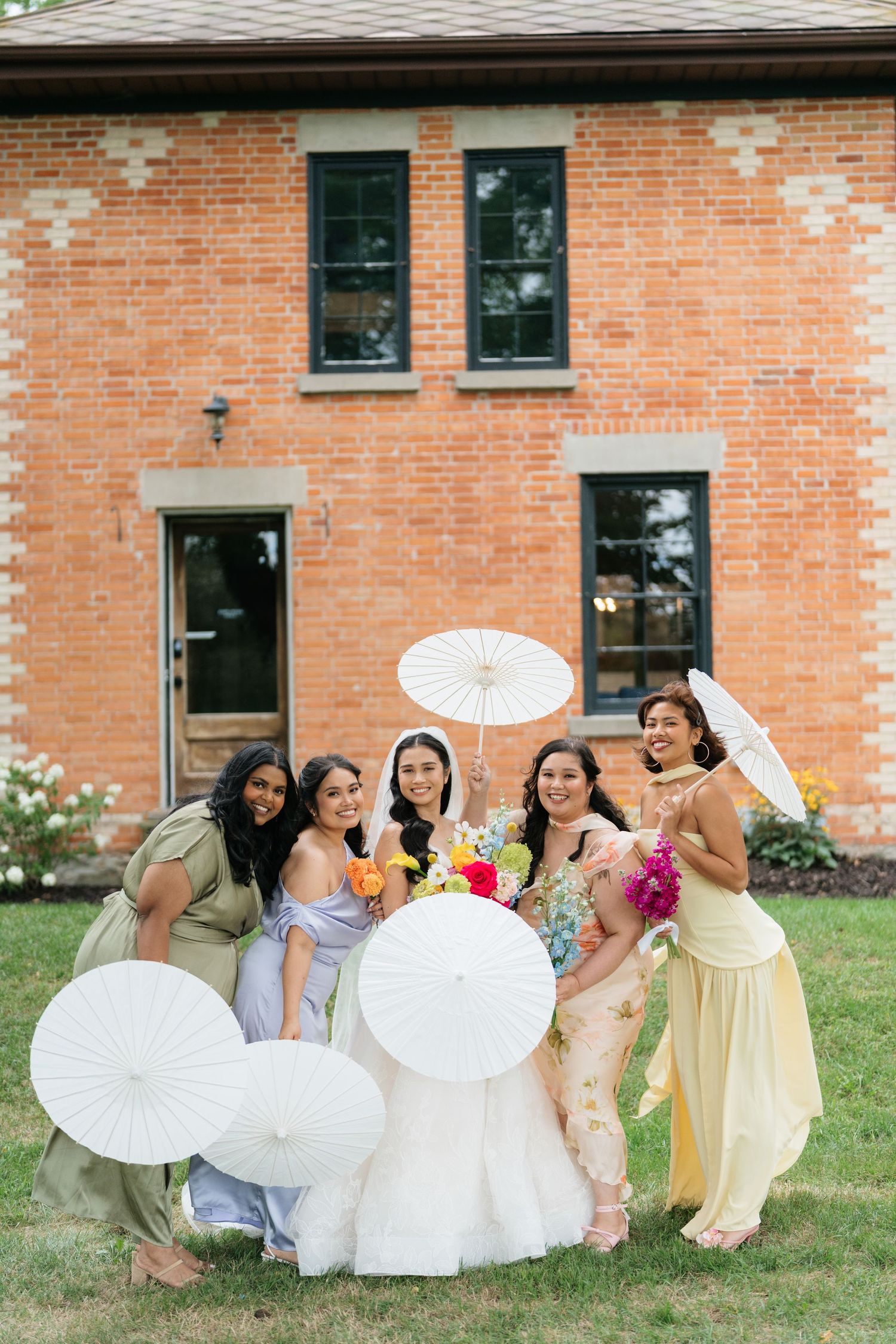 Bridesmaids in pastel dresses pose playfully with white parasols on a lawn in front of a brick building.
