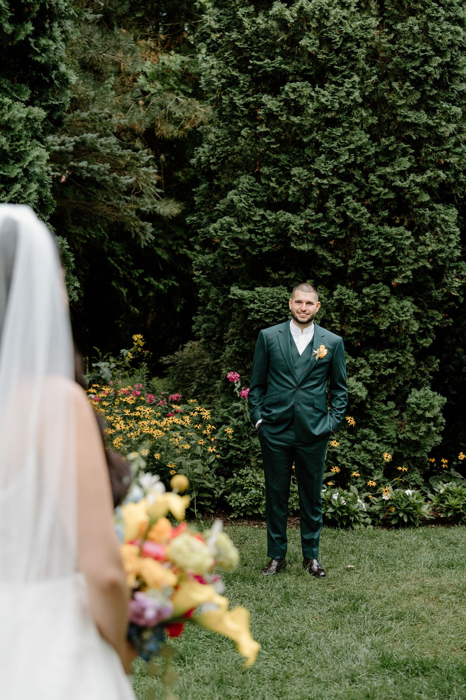 A wedding couple stands separately in a garden setting with green foliage and colorful flowers in the background.
