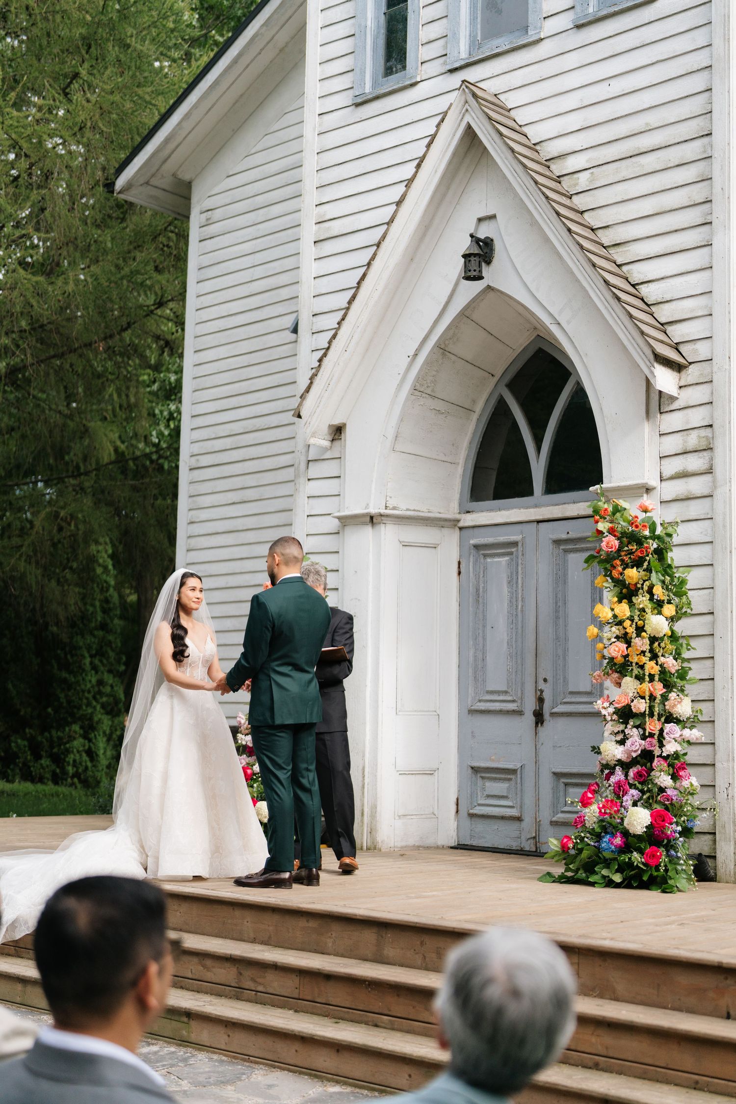 A wedding ceremony takes place at the entrance of a white chapel with an arched doorway and floral decorations.