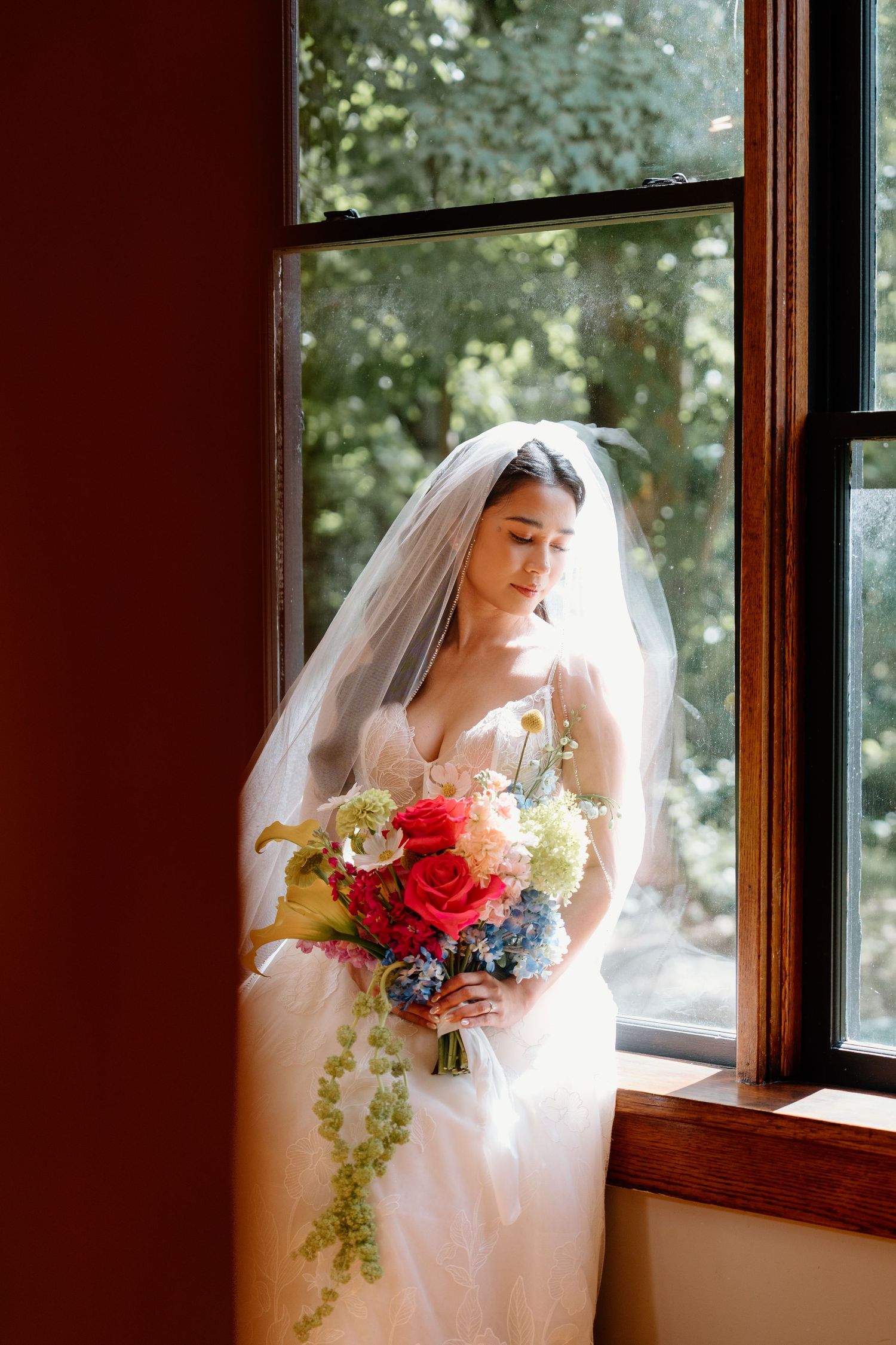 A bride in a white wedding dress and veil holds a vibrant bouquet of red and blue flowers by a sunlit window.
