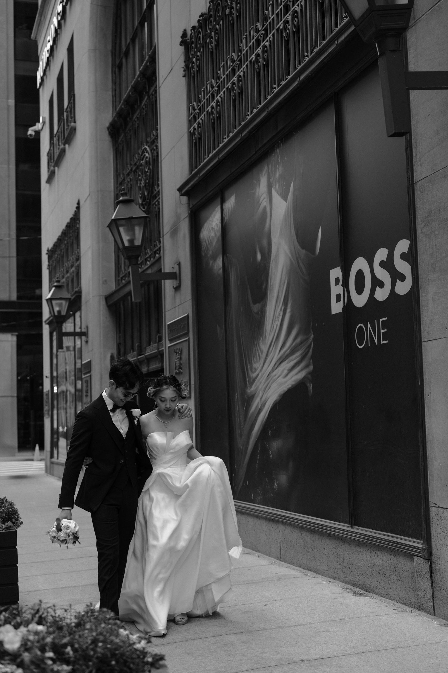 Black and white photo of a couple in wedding attire walking past a Boss store with elegant street lamps in the background.