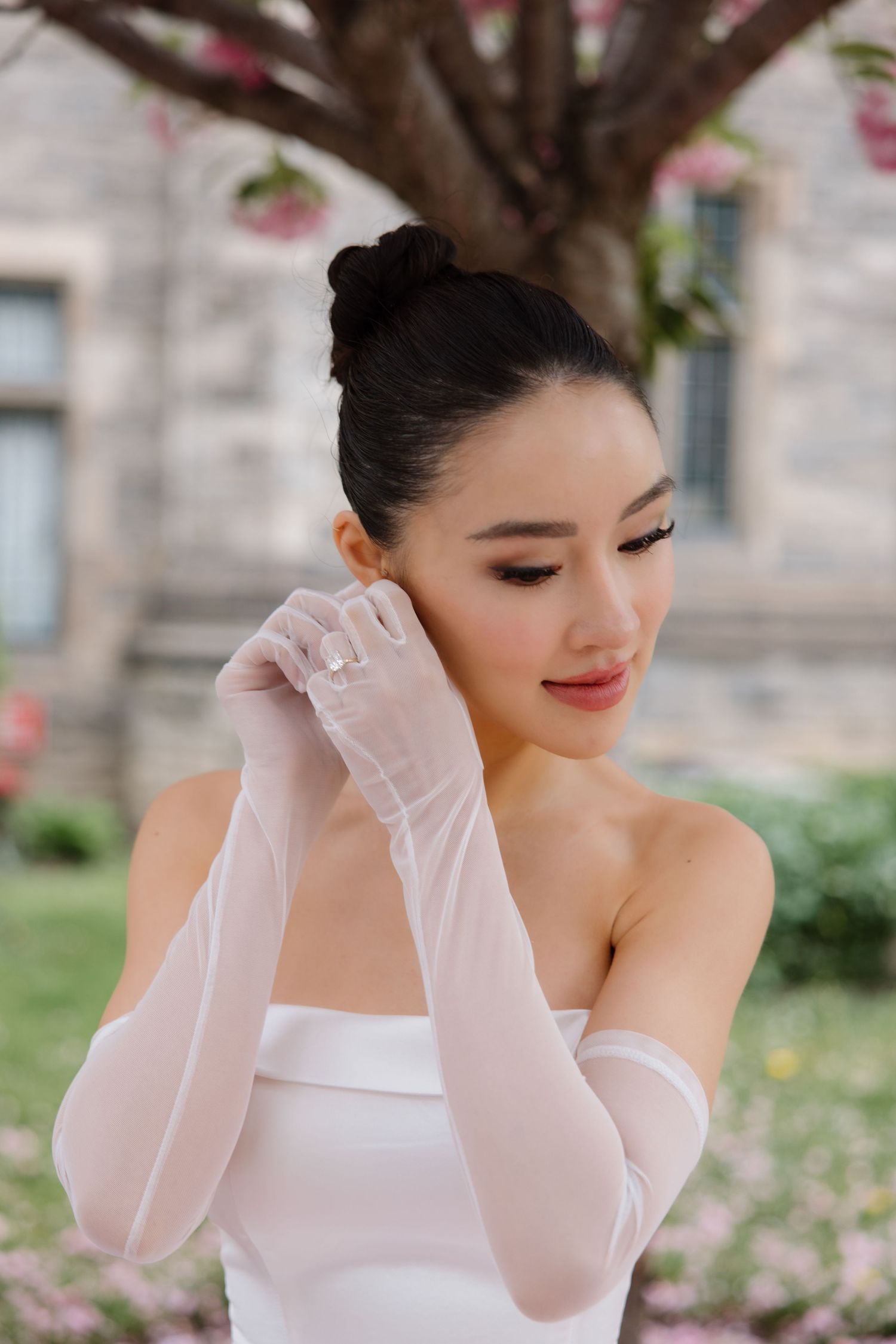 A bride in a white strapless gown and sheer gloves poses gracefully under cherry blossoms in spring.