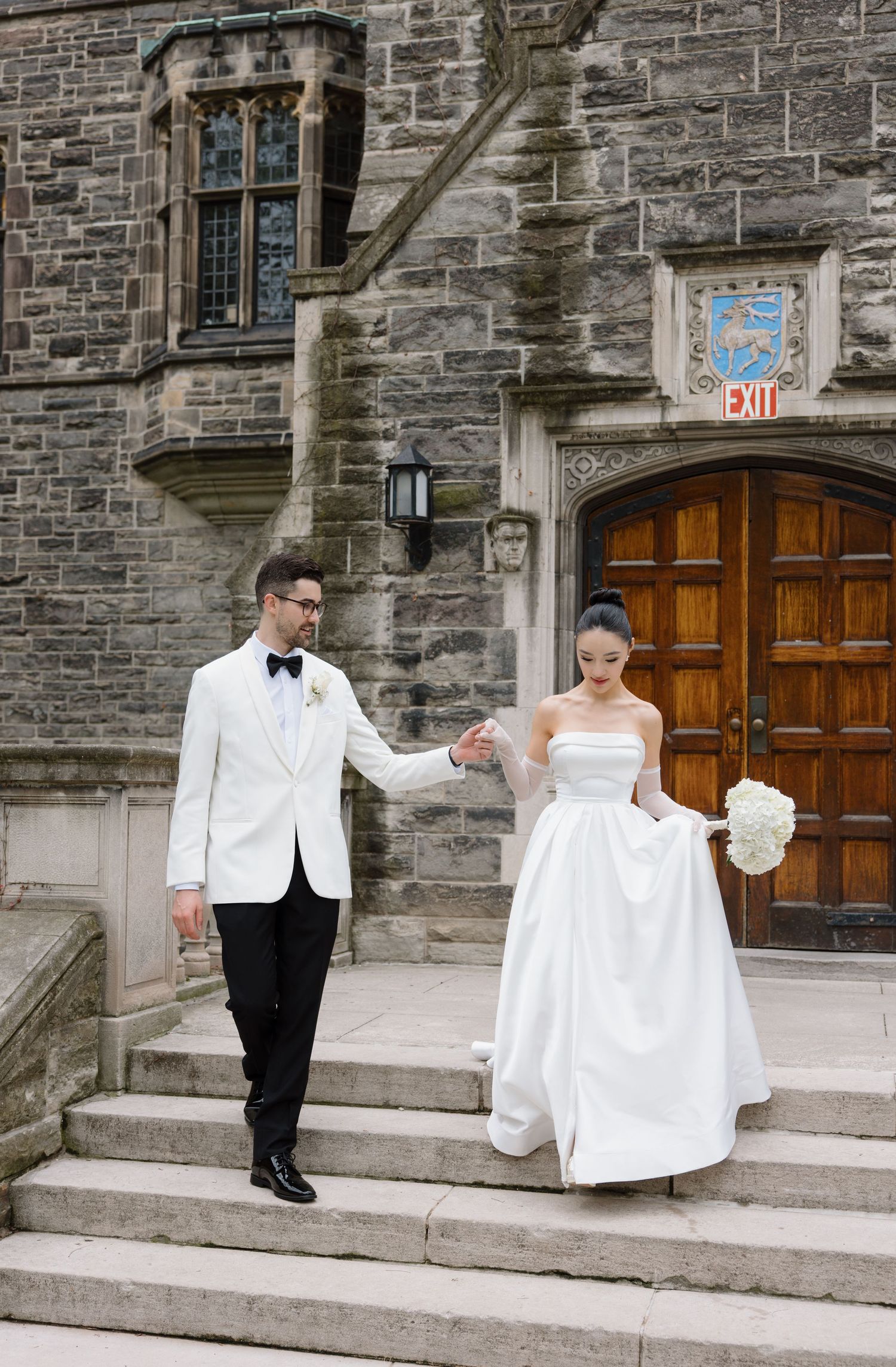 Elegant couple in formal wedding attire descend stone steps of historic Gothic church building with arched wooden door.
