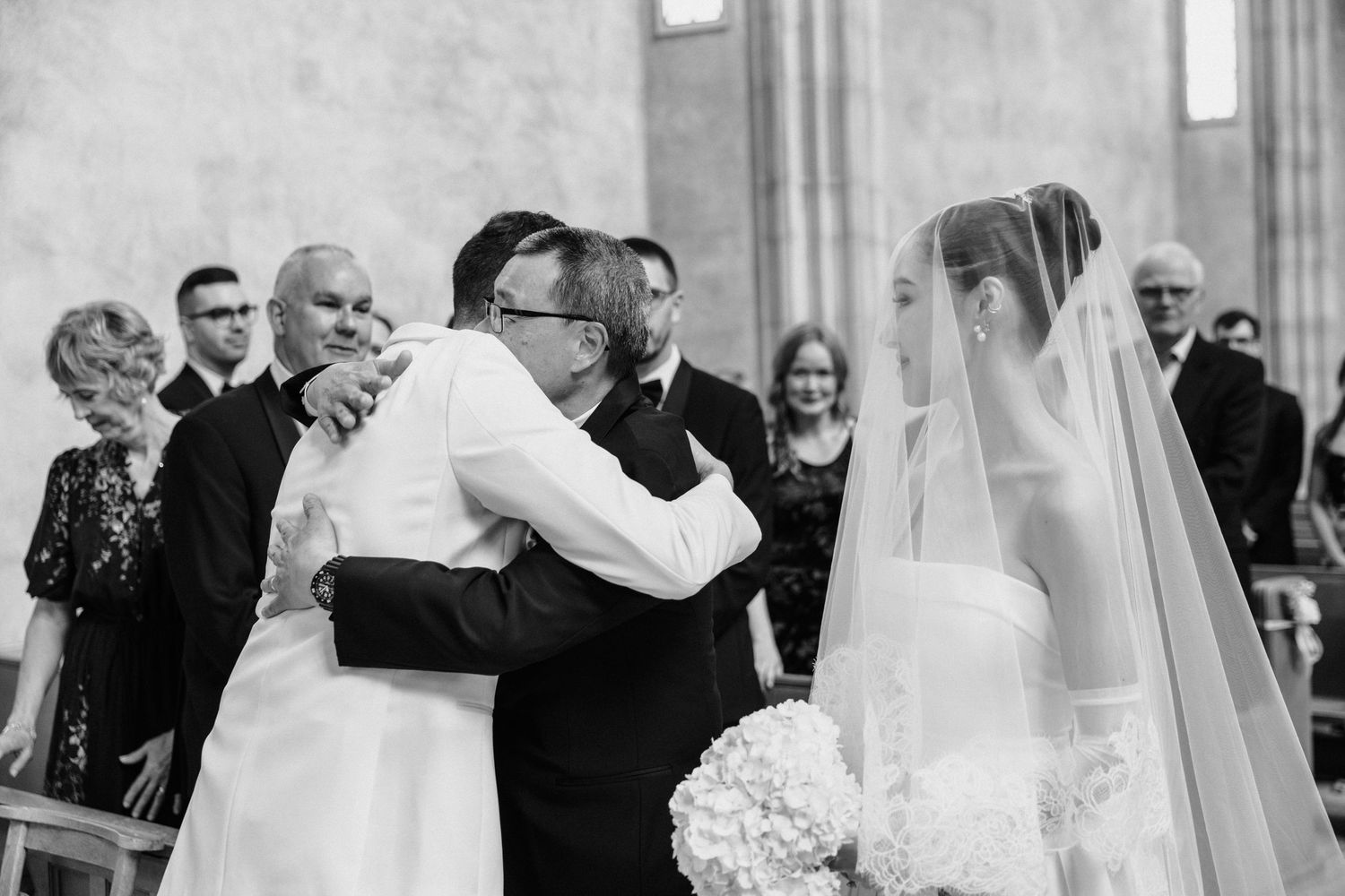 An emotional black and white wedding ceremony moment shows guests embracing as a bride in a veil stands nearby.