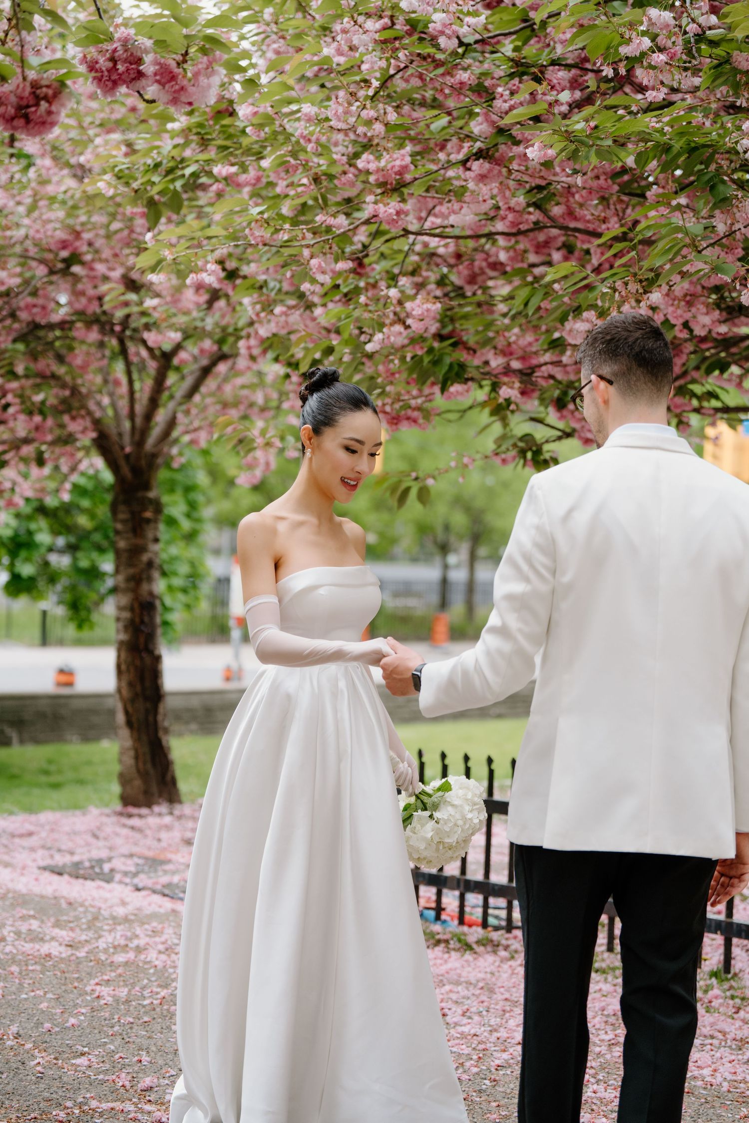 Elegant wedding couple stands beneath blooming cherry blossoms as pink petals fall around them in a spring garden.