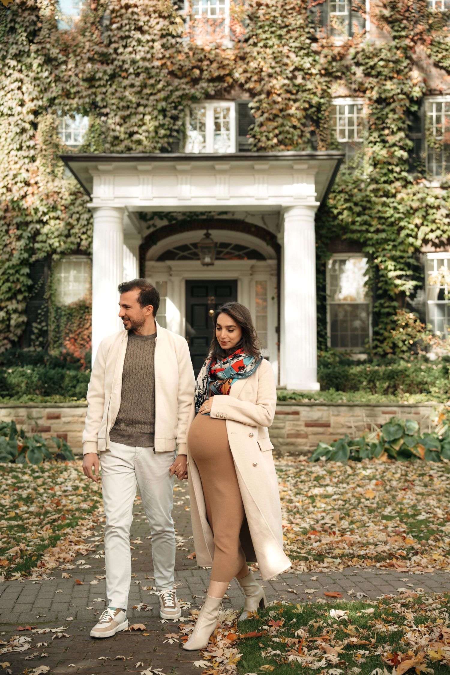 A couple walks together on a leaf-covered pathway in front of an elegant brick building on an autumn day.