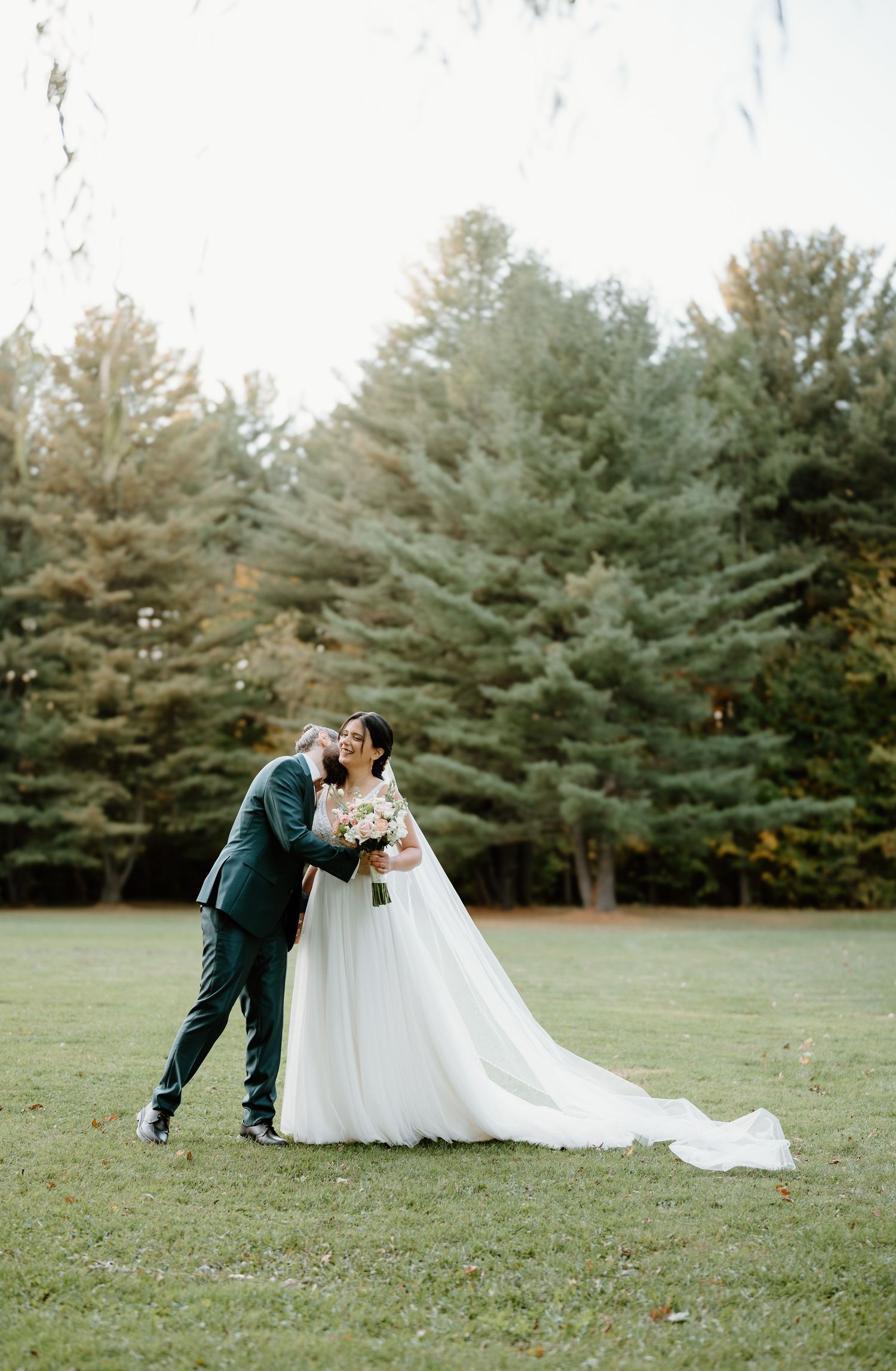 A romantic wedding couple shares a tender moment on a lawn with tall evergreen trees in the background.