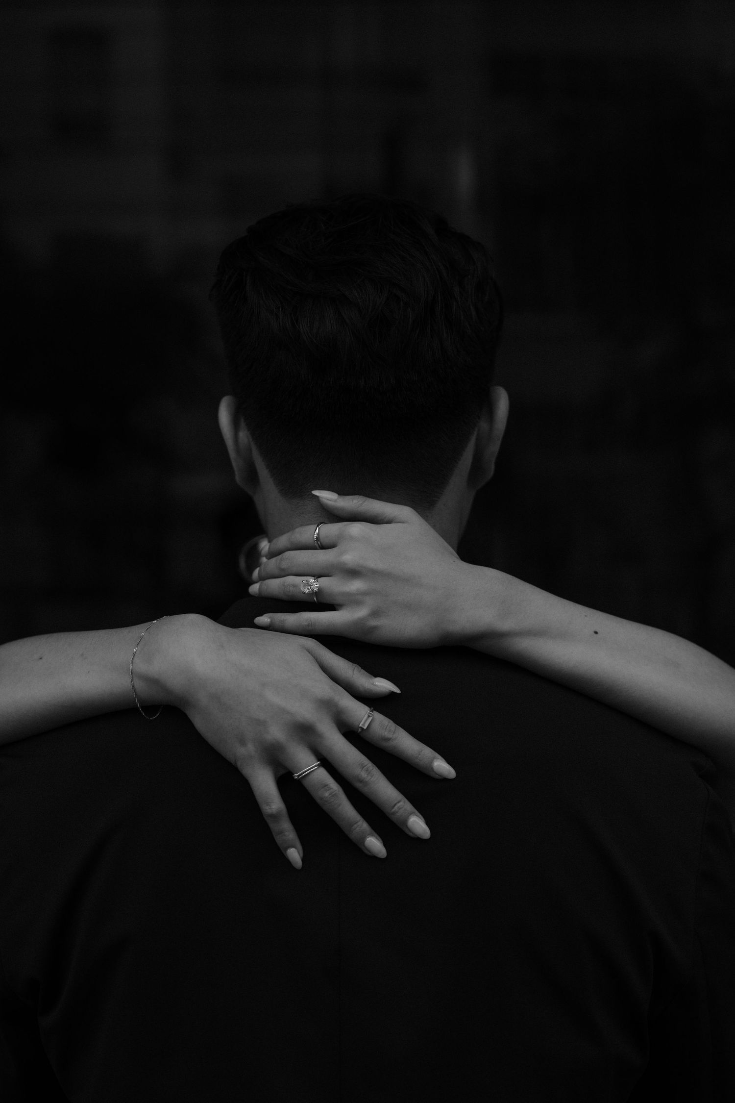 Black and white photo showing hands embracing from behind with one hand on neck and another wrapped around shoulder.