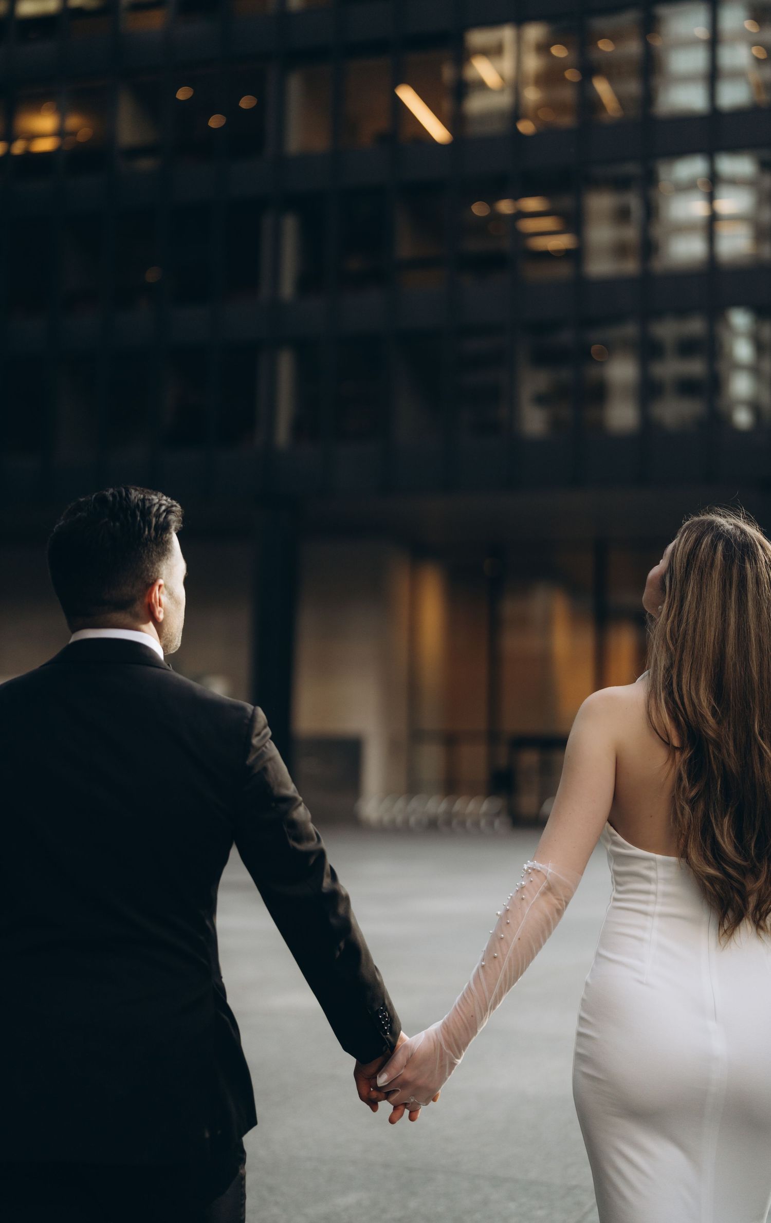 A couple in wedding attire hold hands in front of a modern building at dusk with soft lighting.
