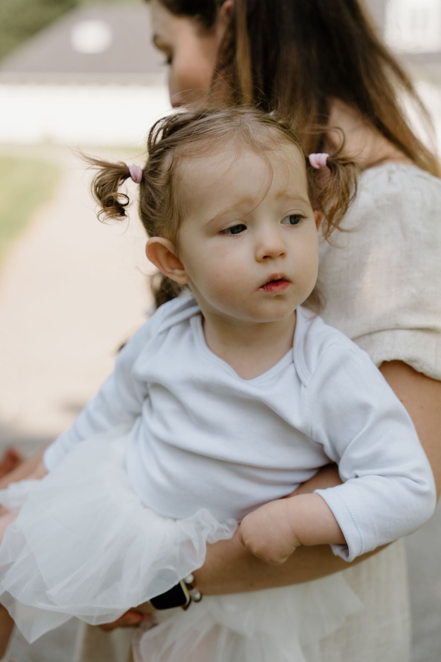 A toddler in a white long-sleeved dress sits on a white surface in a light, airy setting.