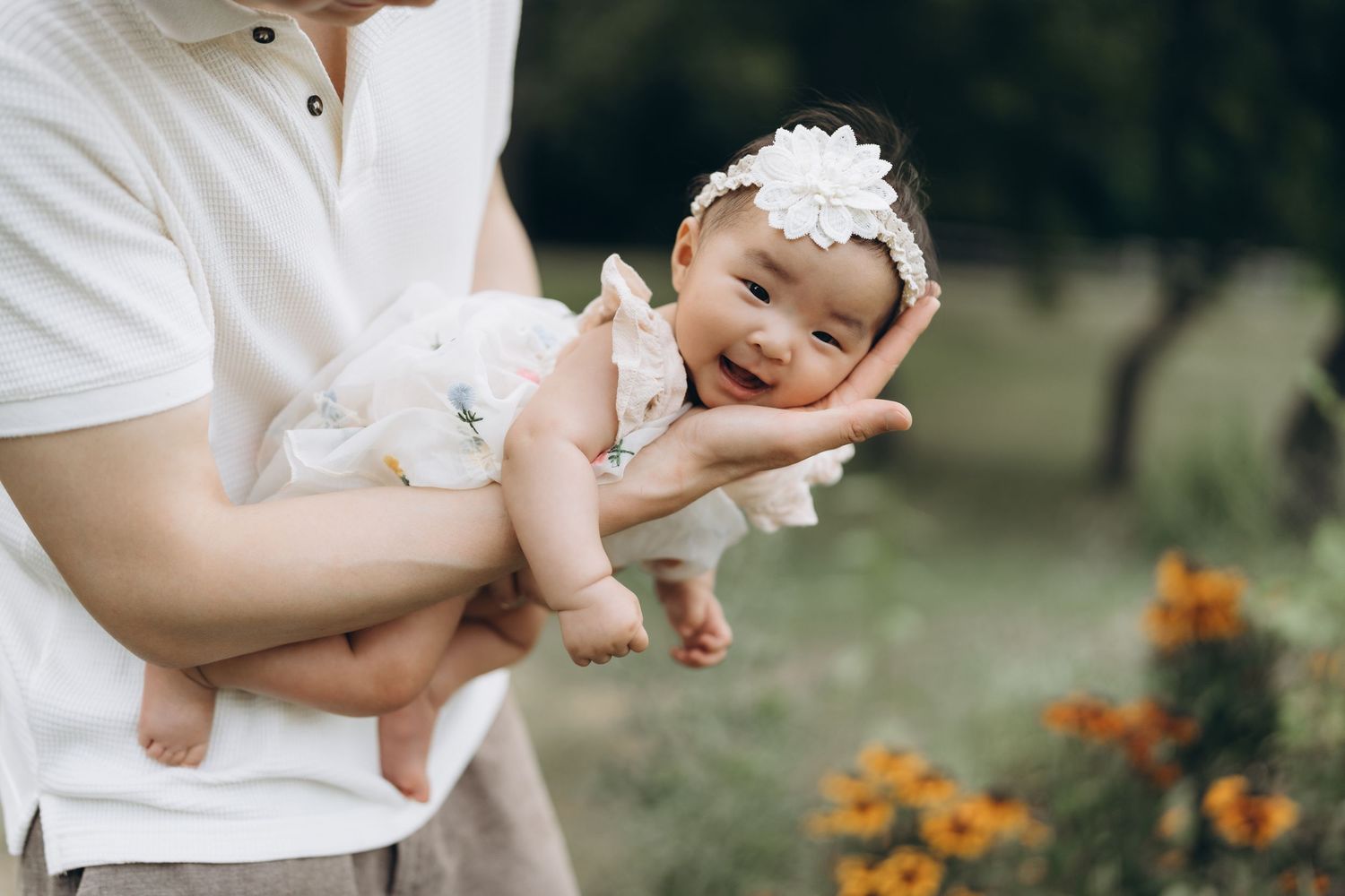 A baby wearing a white headband is held up by adult hands near orange flowers in a garden.