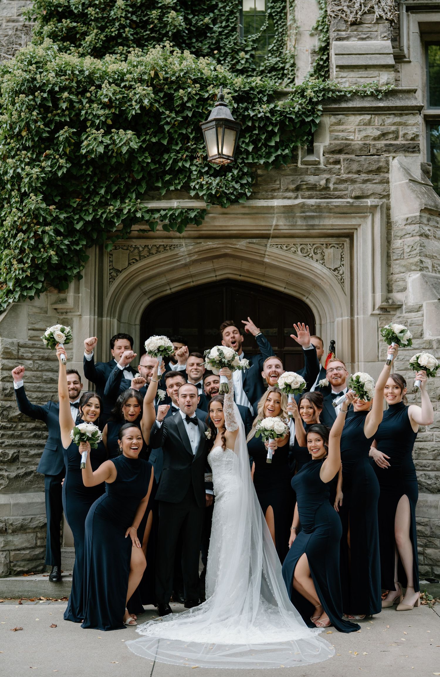 Wedding party in navy blue dresses and suits poses together in front of stone archway with ivy growing above.