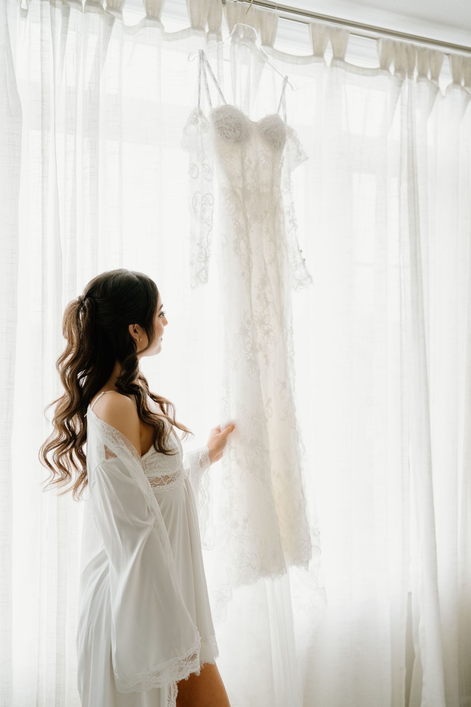 Ethereal silhouette in white stands by sheer curtains in morning light creating a dreamy boudoir atmosphere.