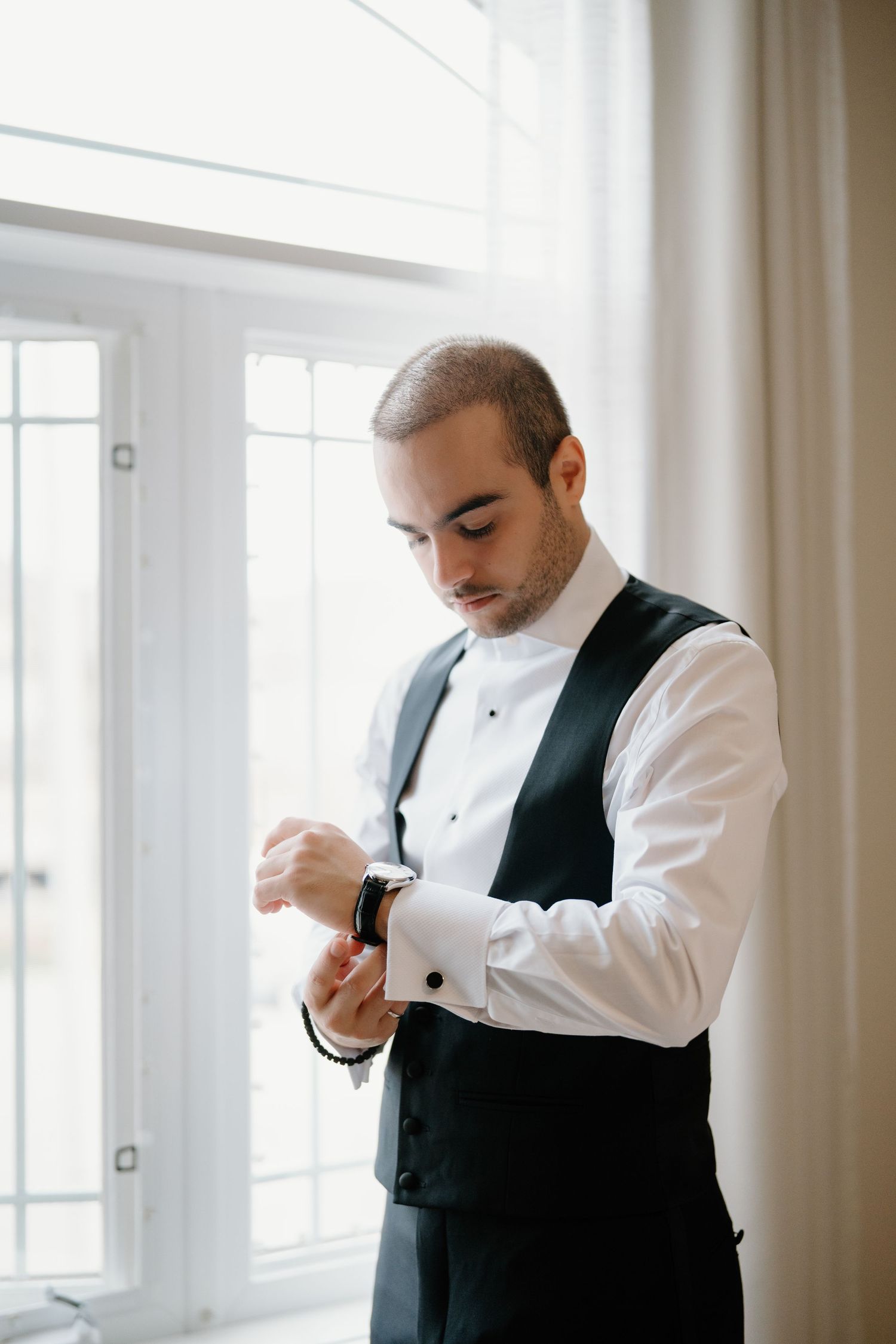 A person in a white shirt and black vest adjusts their attire in a bright room with windows.