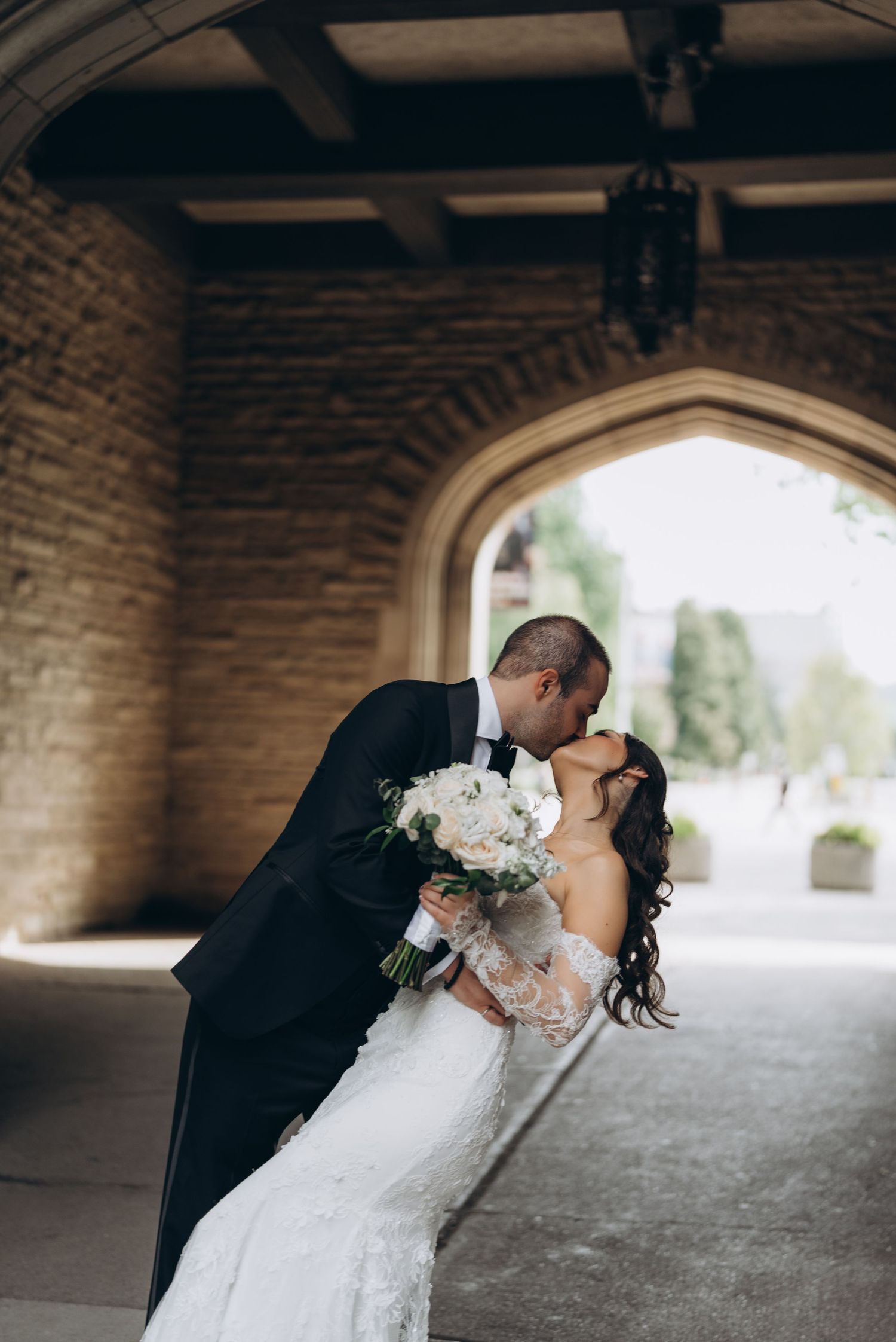 A romantic wedding couple shares a kiss under a stone archway while the bride holds a white floral bouquet.