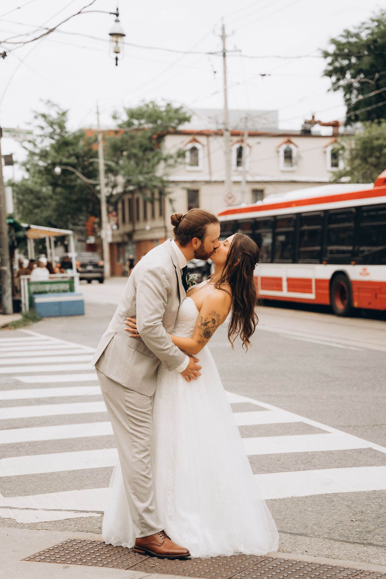 A couple in wedding attire dances playfully on a crosswalk with a streetcar visible in the urban background.