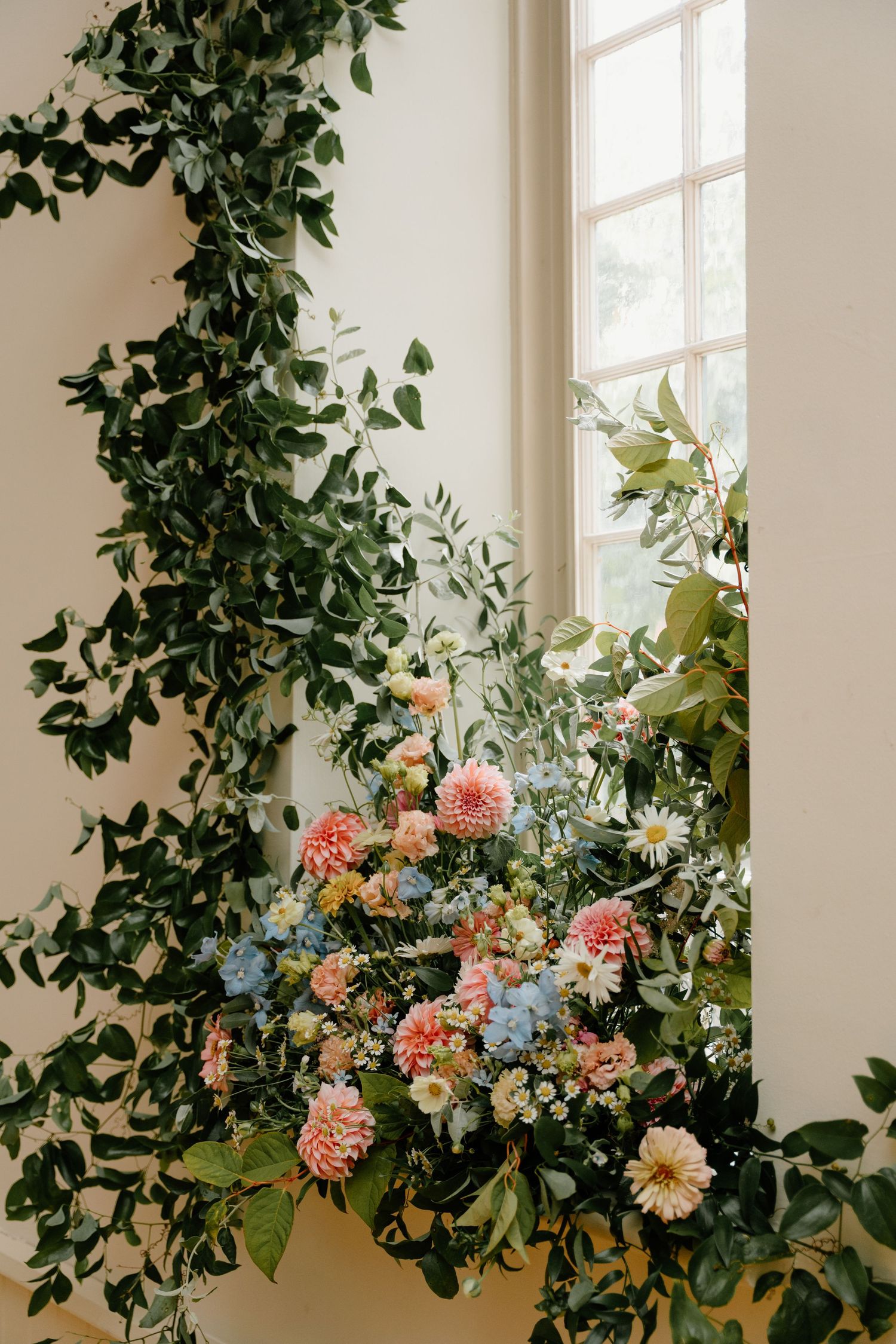 Colorful floral arrangement with pink and blue blooms adorns a bright interior space with climbing green vines near windows.