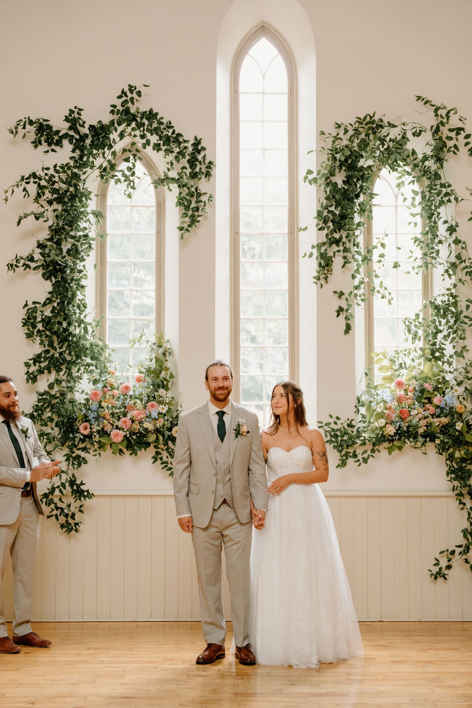 A couple shares a romantic dance in a bright white chapel room decorated with climbing greenery and arched windows.