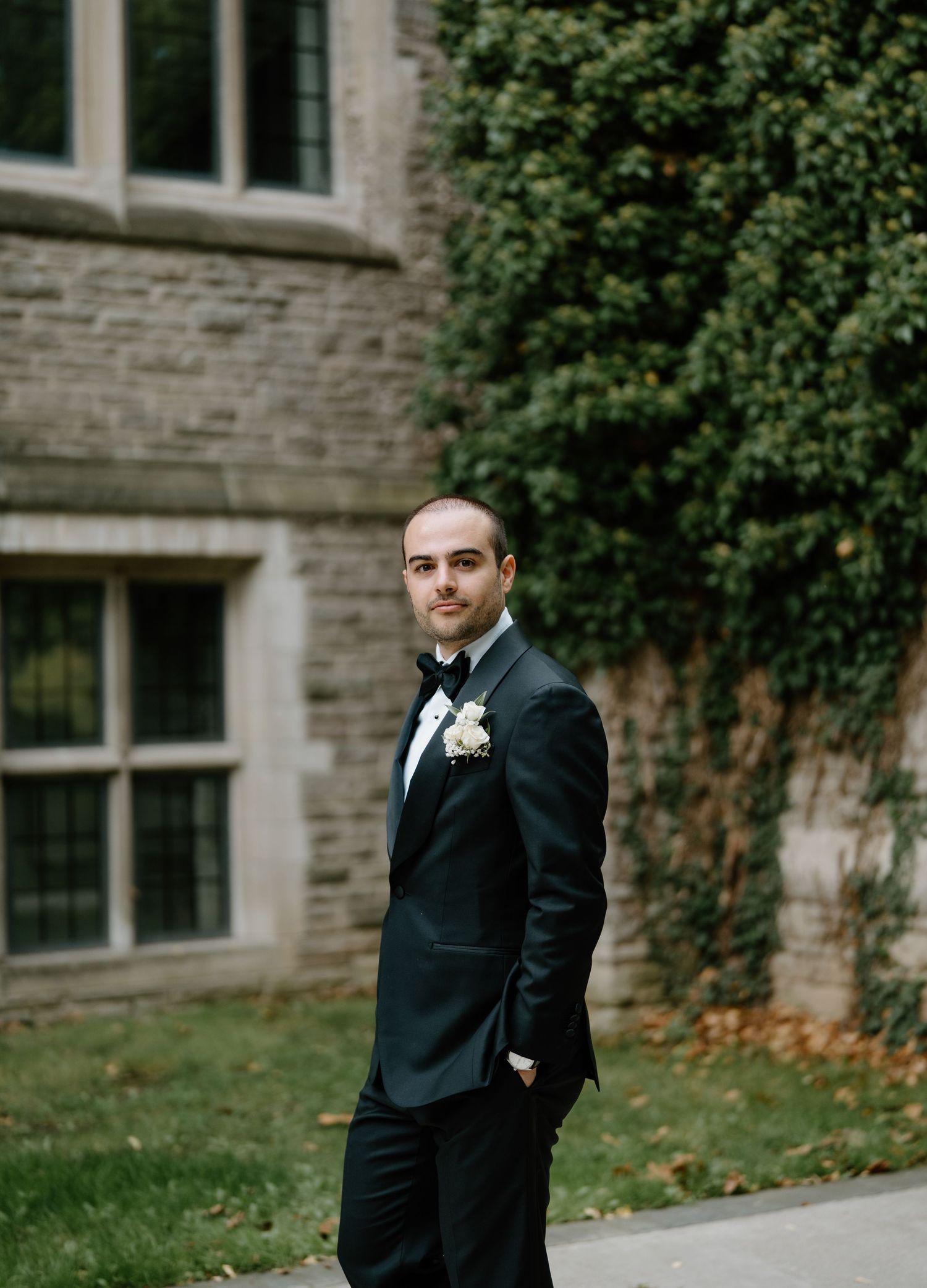 A groom stands confidently in formal attire on the grounds of a historic stone building.