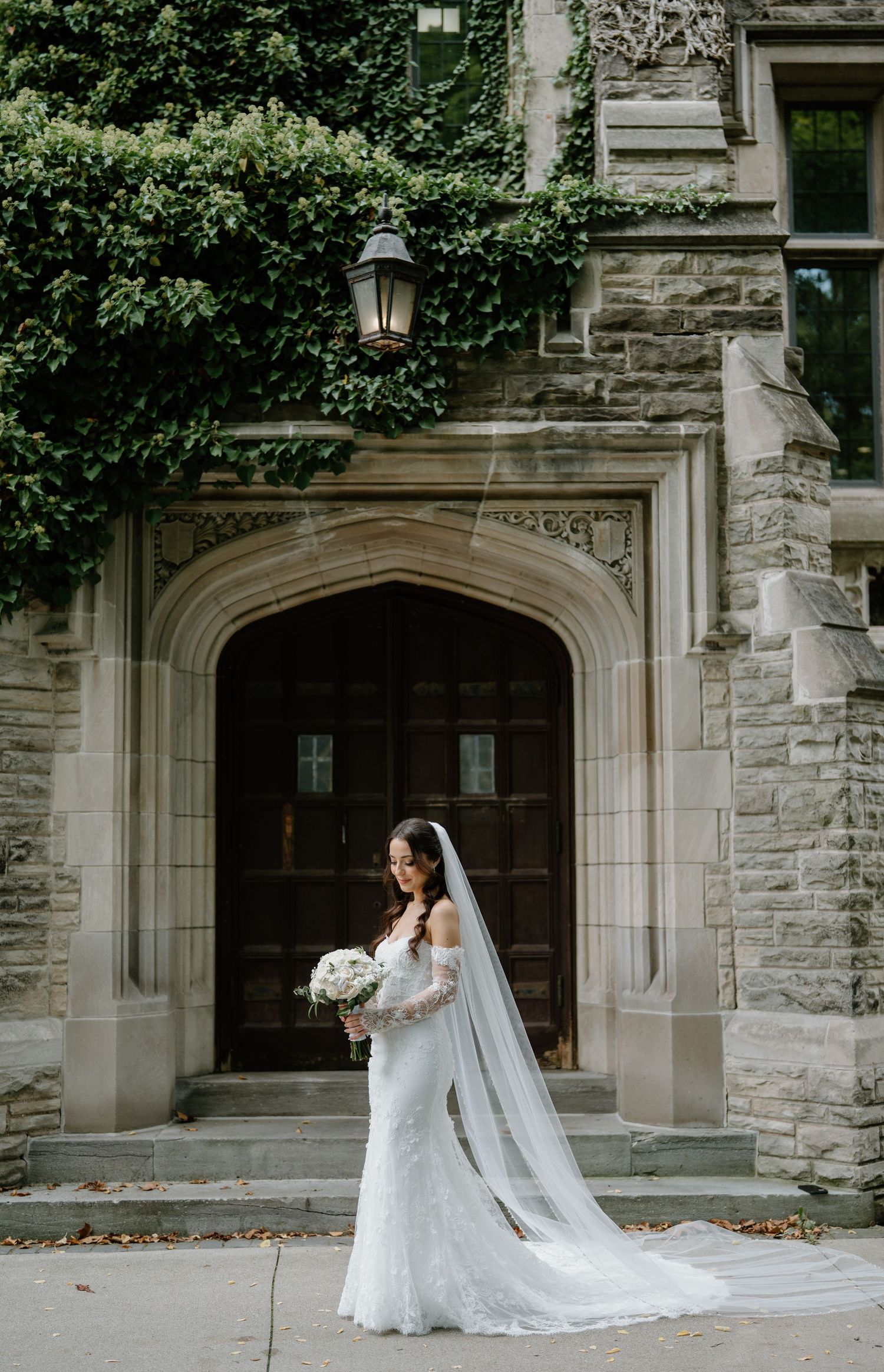 A bride in a flowing white gown and veil poses elegantly against a stone building with gothic architectural details.