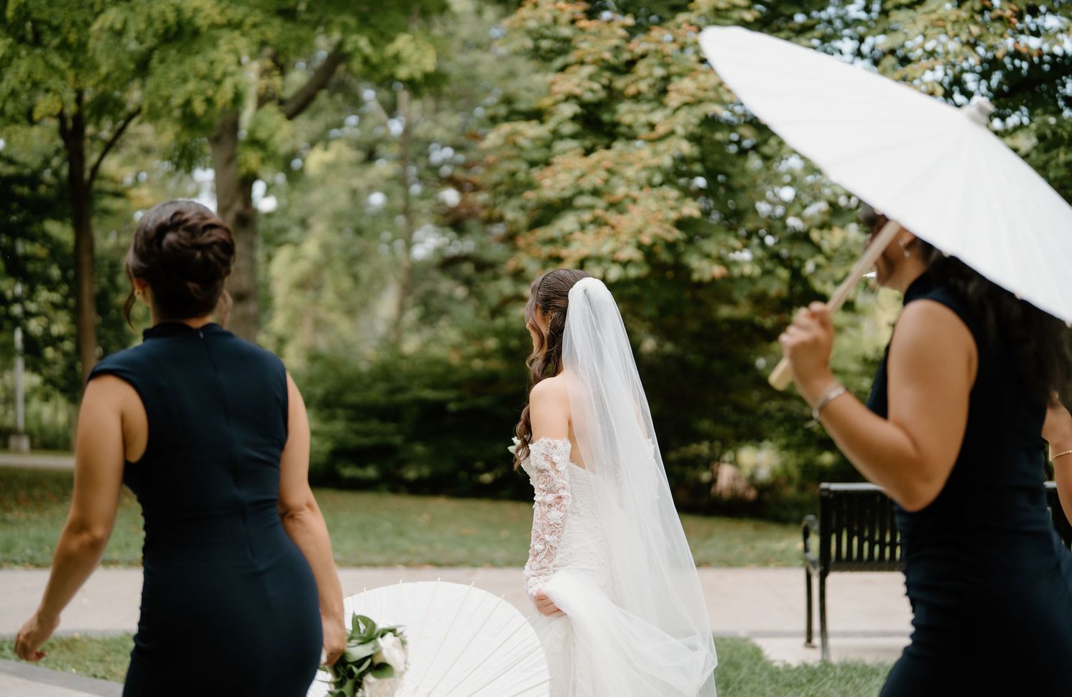 Bridesmaids in black dresses walk with white parasols through a lush garden path.