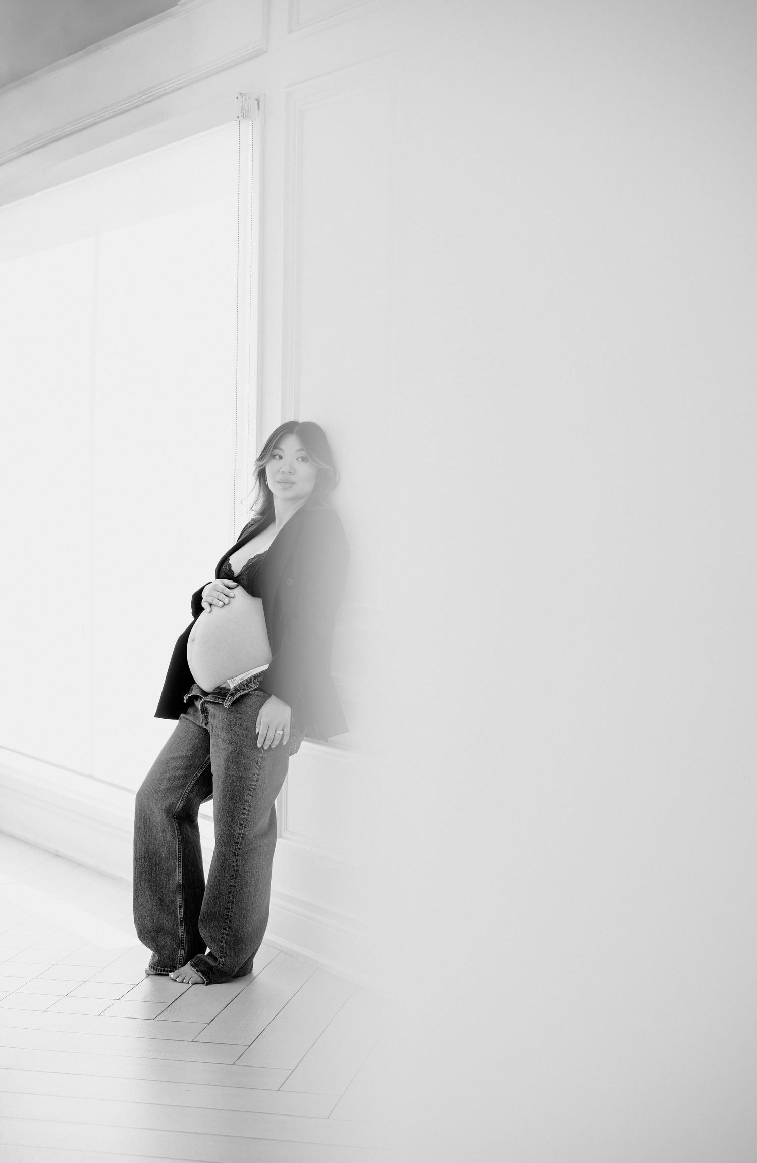 A person in dark clothing poses artistically against a stark white wall and floor in a minimalist setting.