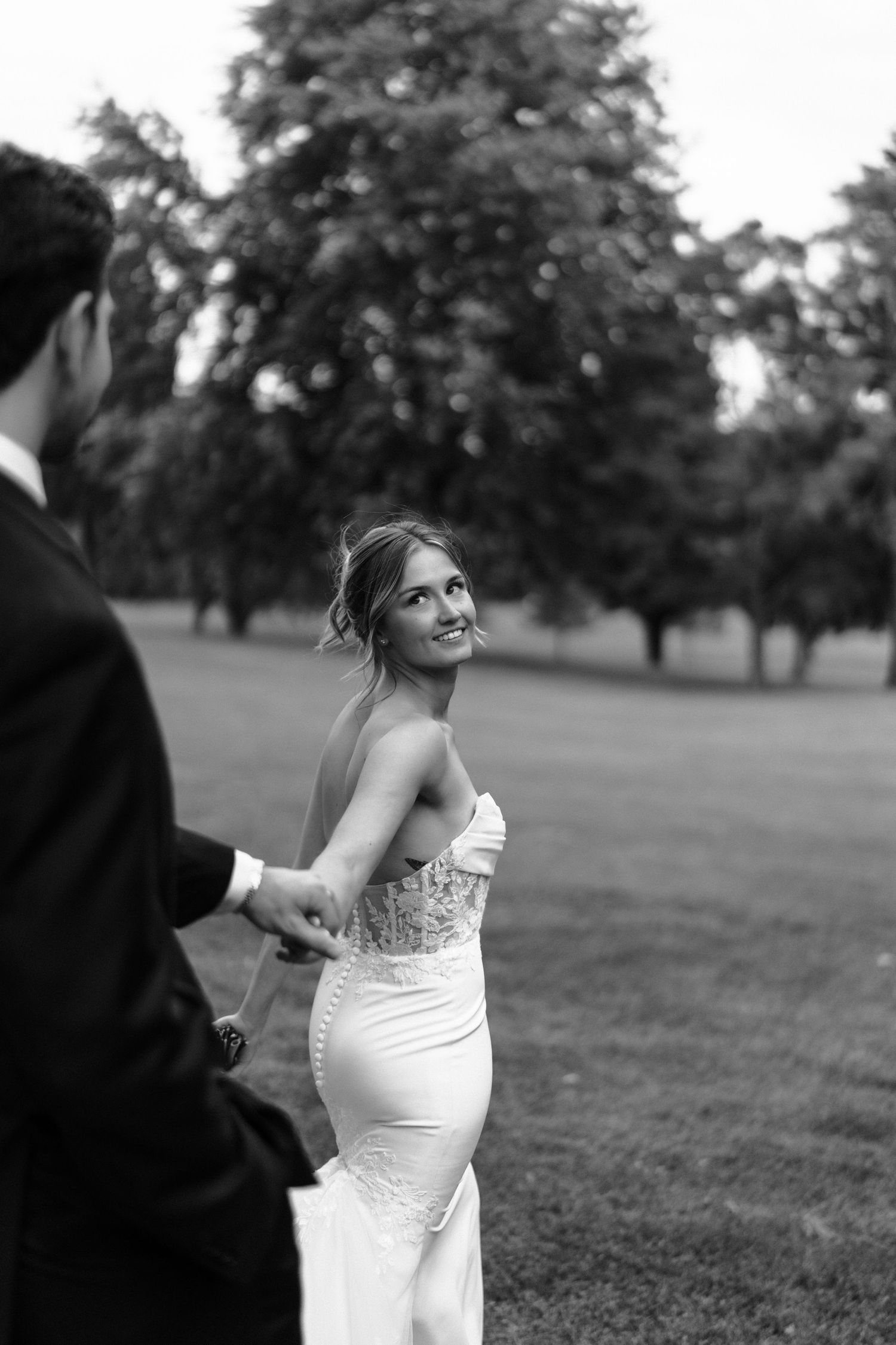 A bride in a white strapless wedding dress smiles joyfully while standing in a grassy outdoor setting.