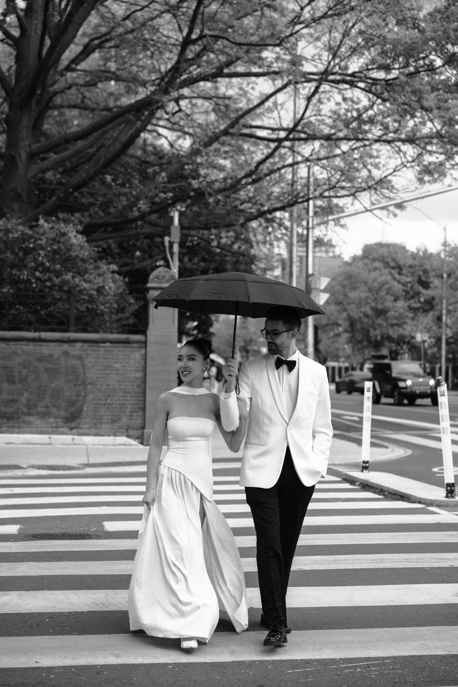 A couple in wedding attire walks across a crosswalk sharing a black umbrella on an overcast day.