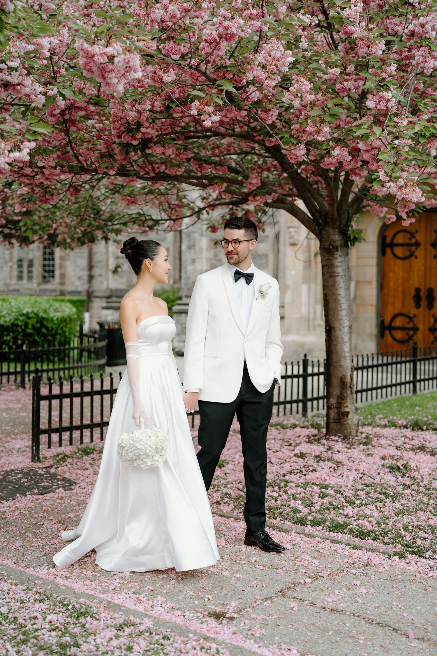 A springtime wedding portrait under pink cherry blossoms with a white dress and white tuxedo jacket.