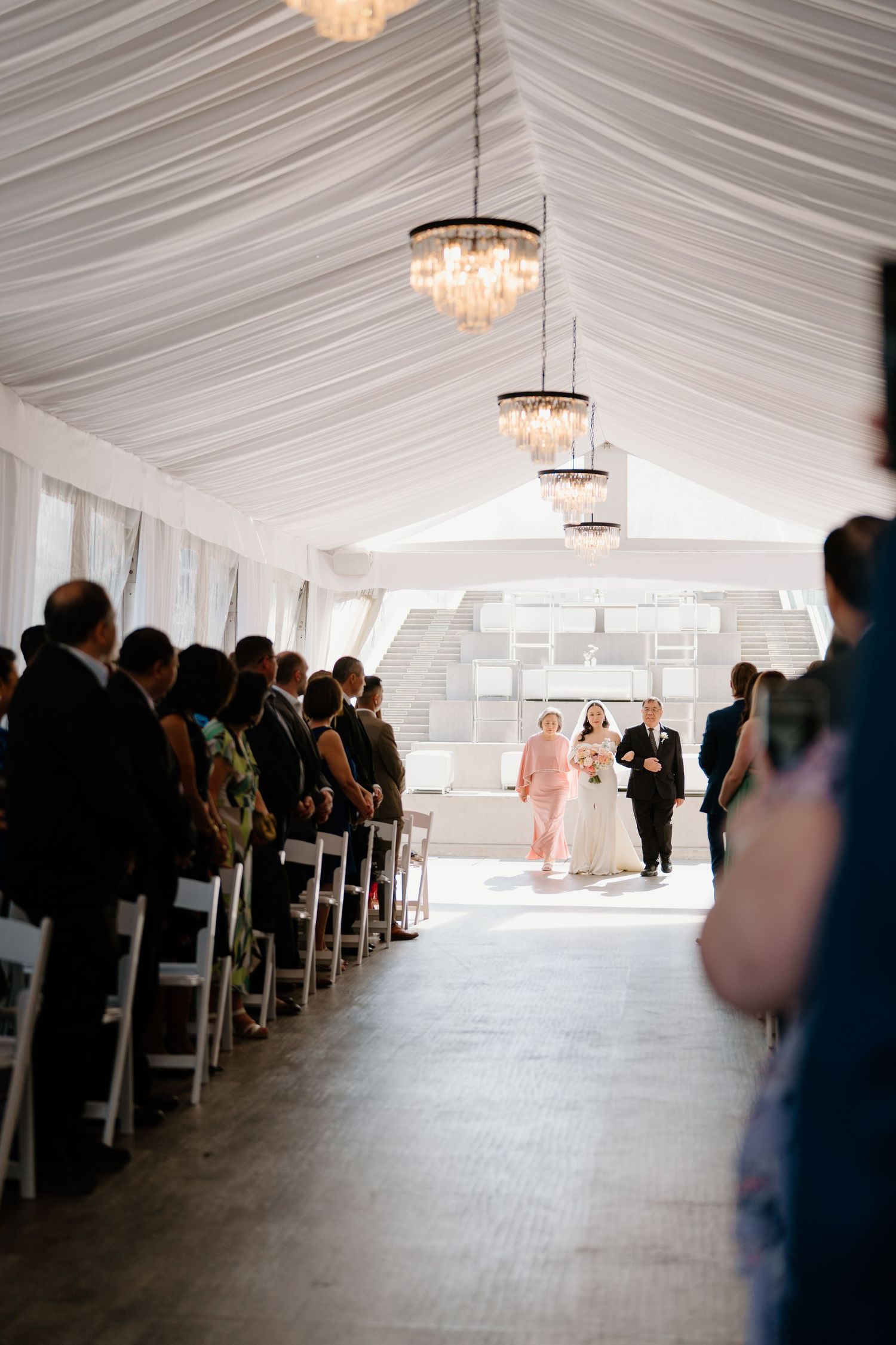 Indoor wedding ceremony in a white draped tent venue with elegant crystal chandeliers and guests seated in rows.