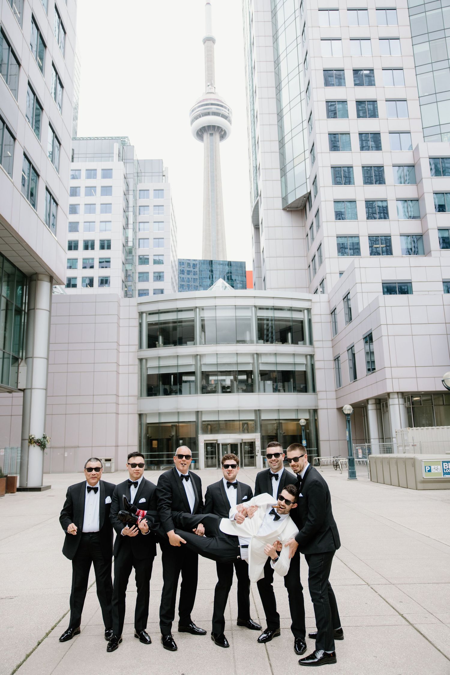 Group in formal black suits pose in front of white buildings with Toronto CN Tower visible in background.