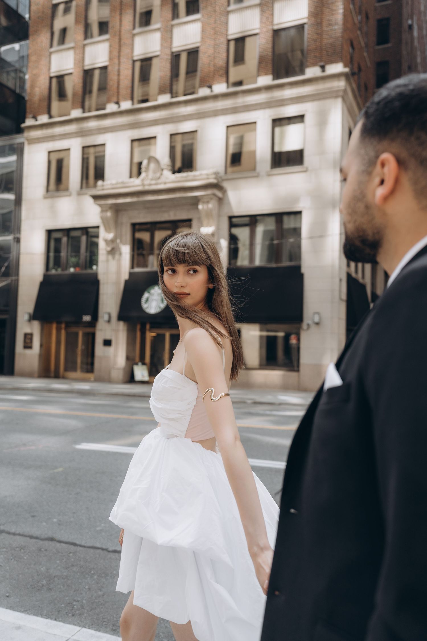 Woman in flowing white dress walking past elegant historic building facades in urban setting.