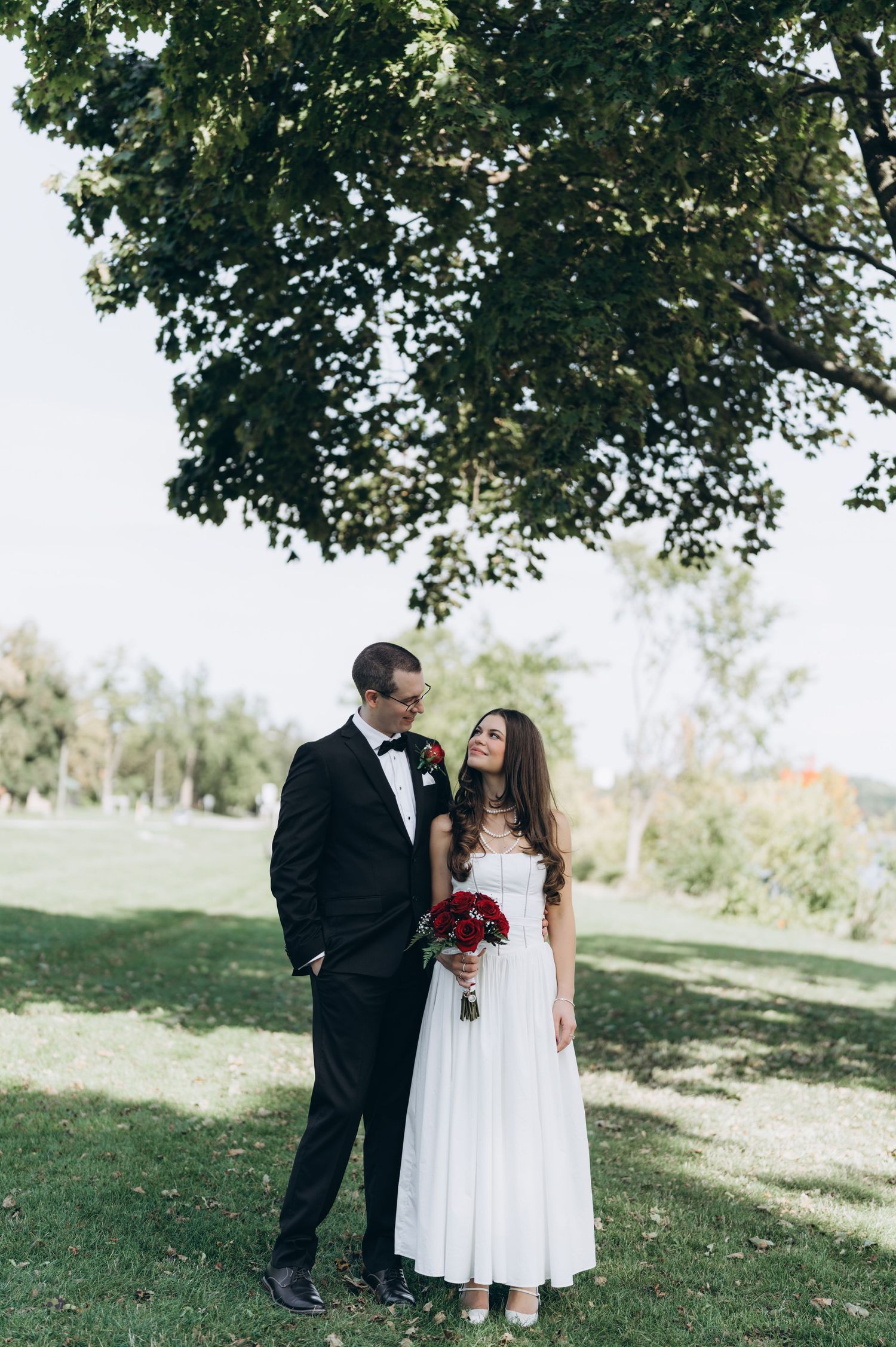 A bride in white dress holds red bouquet while standing with groom in black suit under green tree branches in outdoor setting.