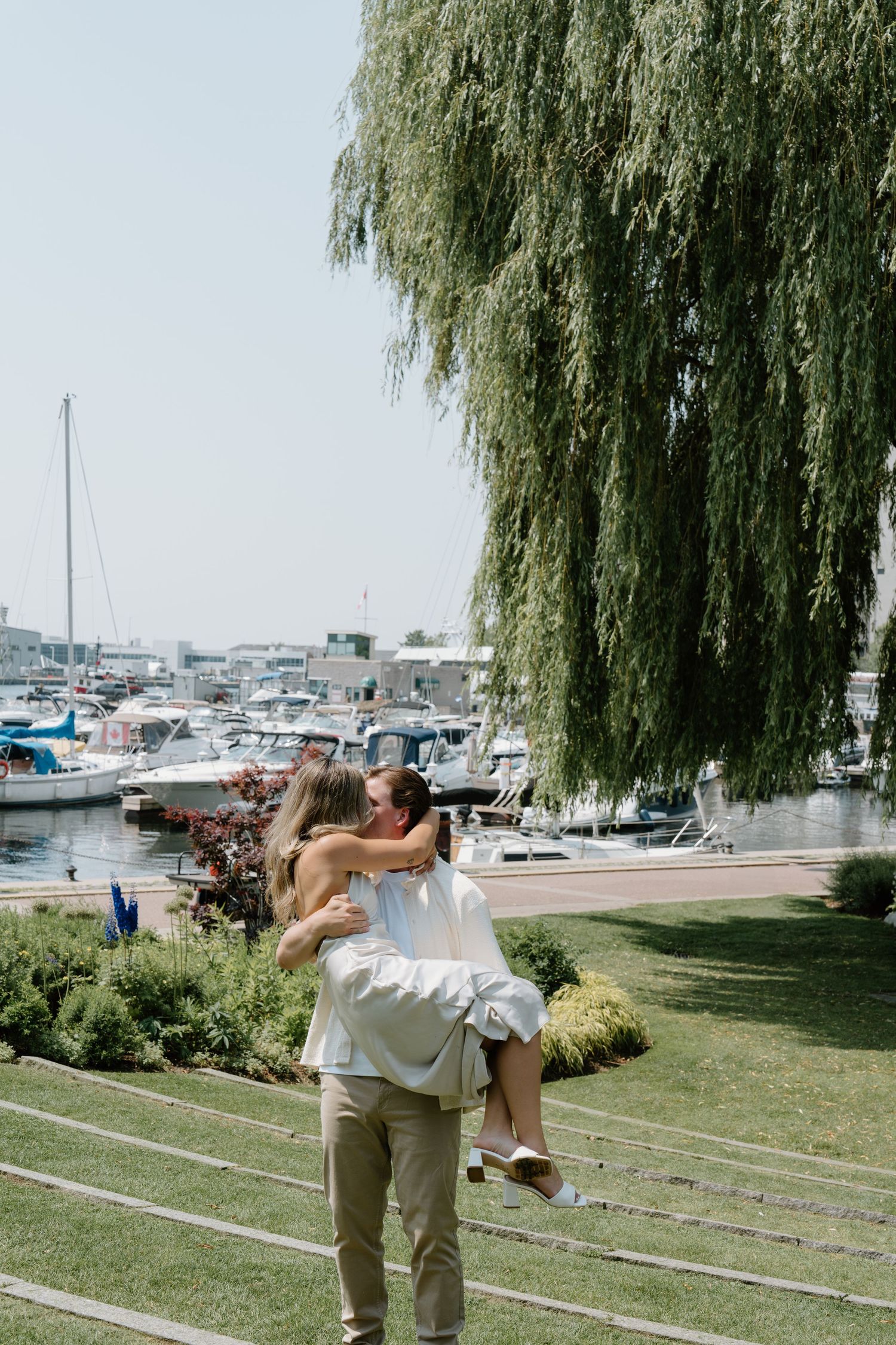 A romantic couple dances and dips near a marina with boats and willow trees in the background.