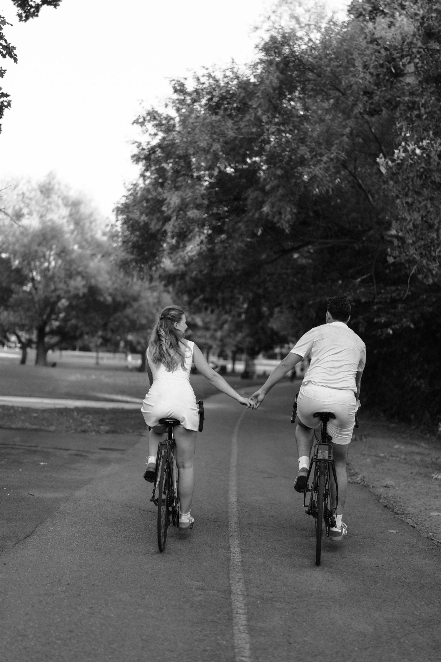 Two cyclists riding bikes together down a tree-lined path in black and white photography.