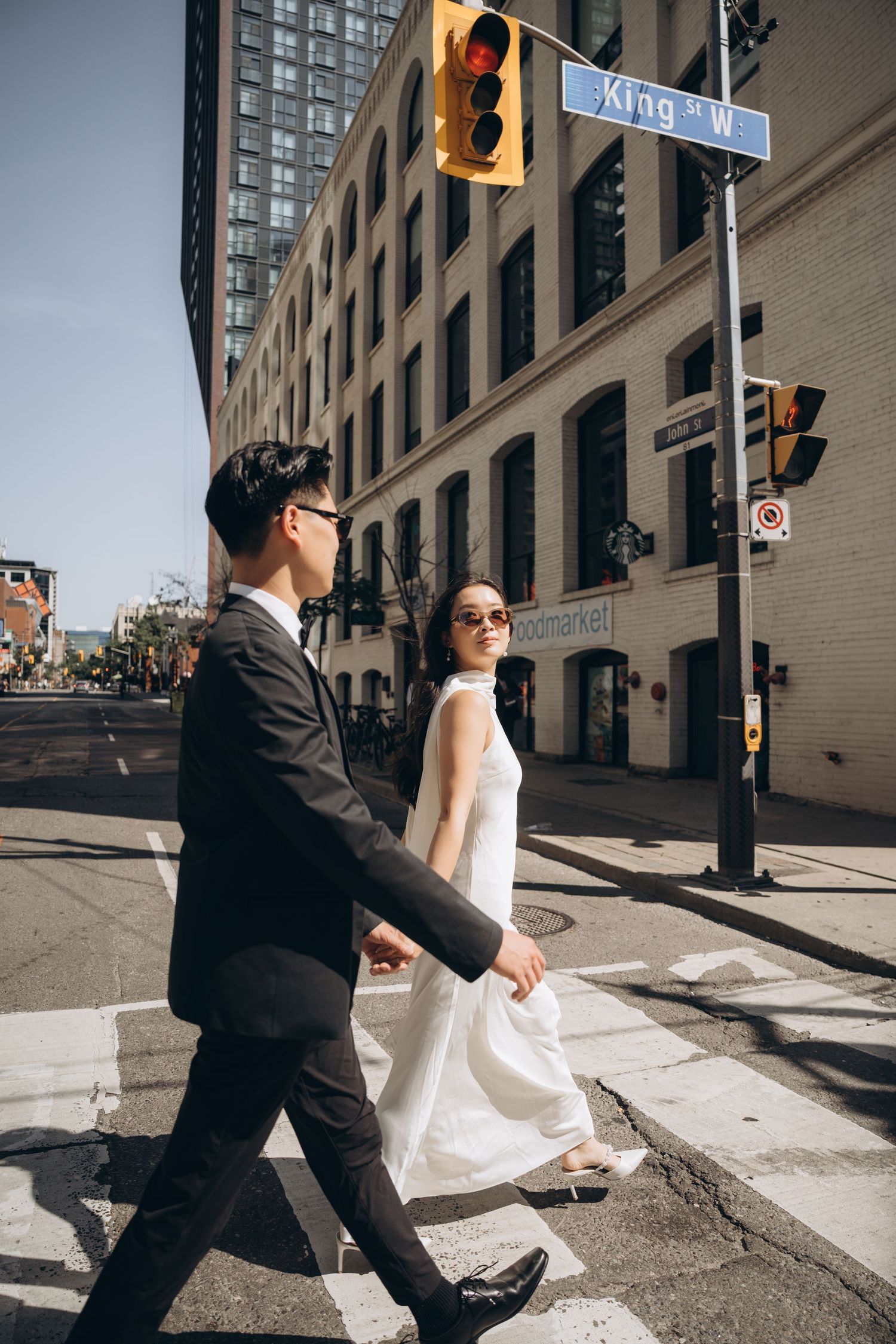 A couple in wedding attire performs dynamic dance moves on a sunny city street corner.