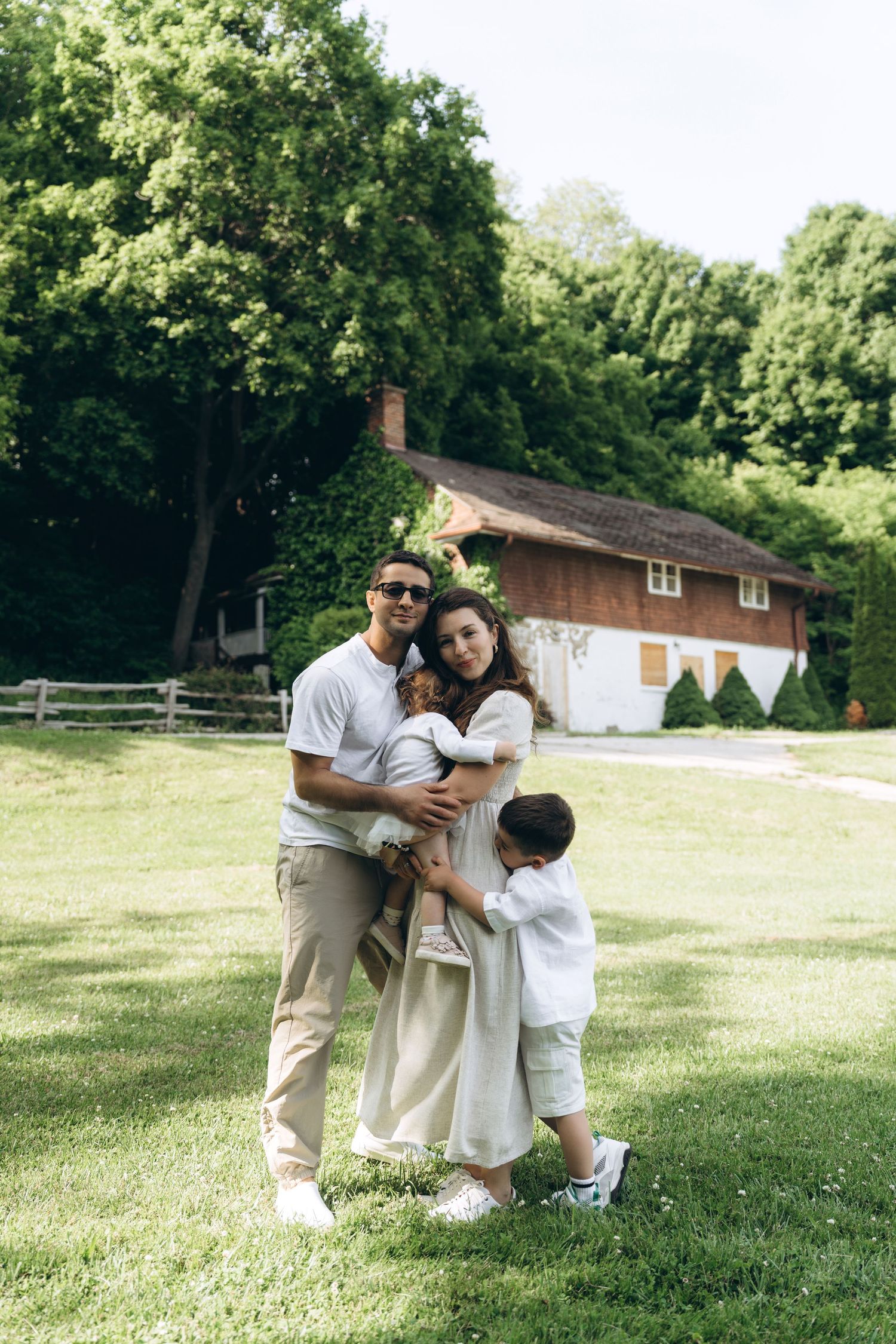 A family stands together on a sunny lawn in front of a brown house surrounded by green trees.